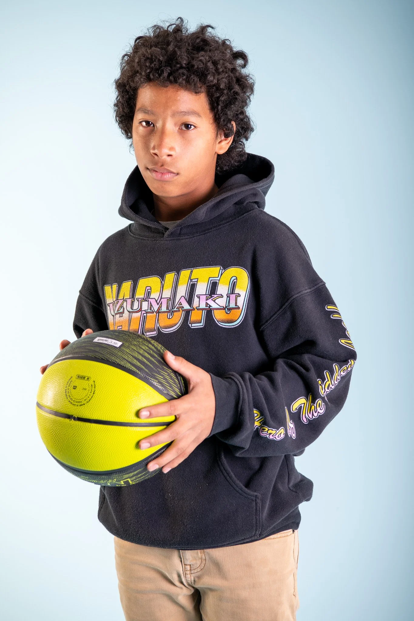 A young boy with curly hair holding a yellow and black basketball, wearing a black hoodie with colorful text and beige pants, standing against a light blue background.