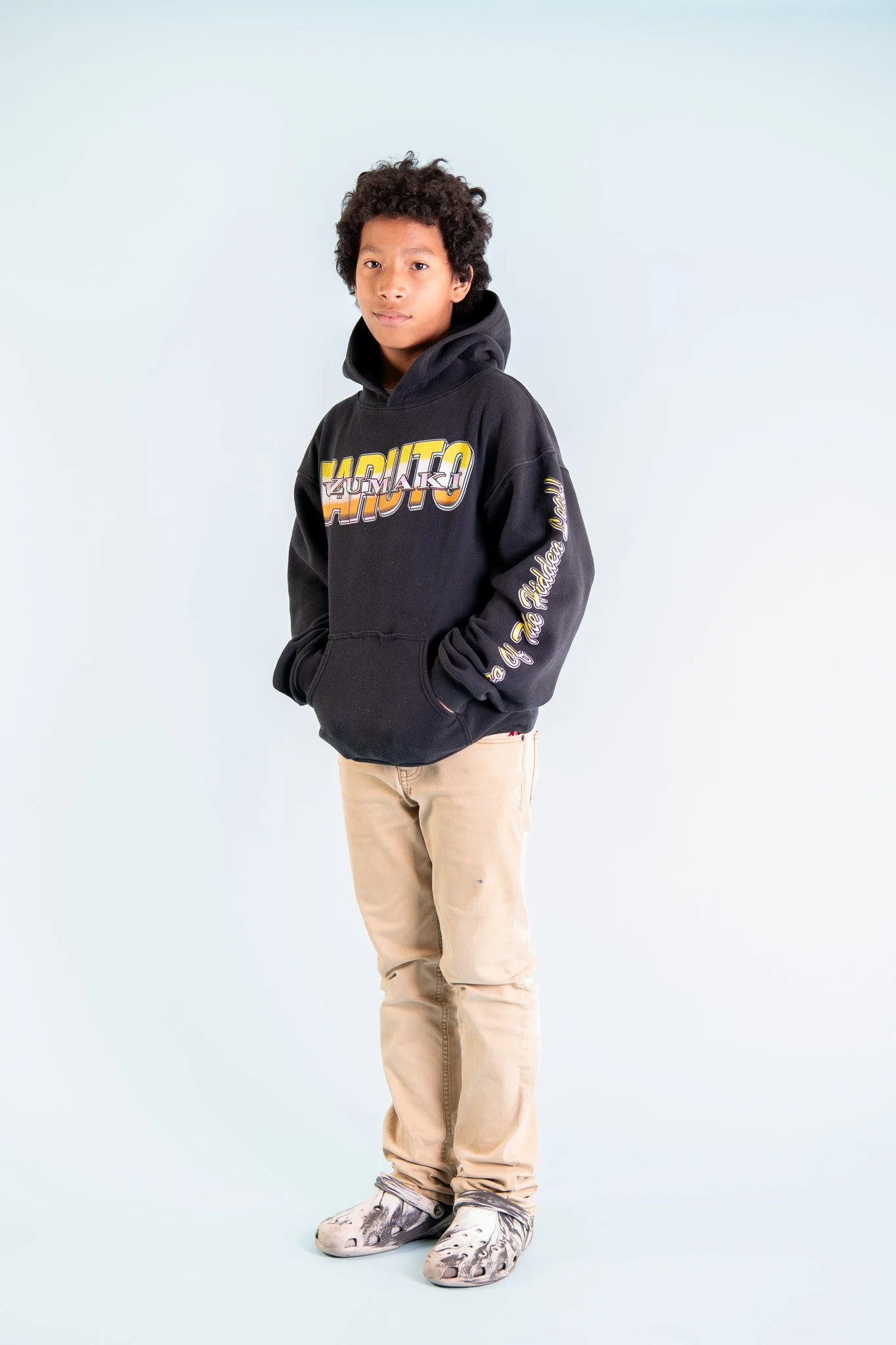 A young boy with curly hair standing against a light background, wearing a black hoodie with colorful graphic text, beige pants, and patterned sneakers.