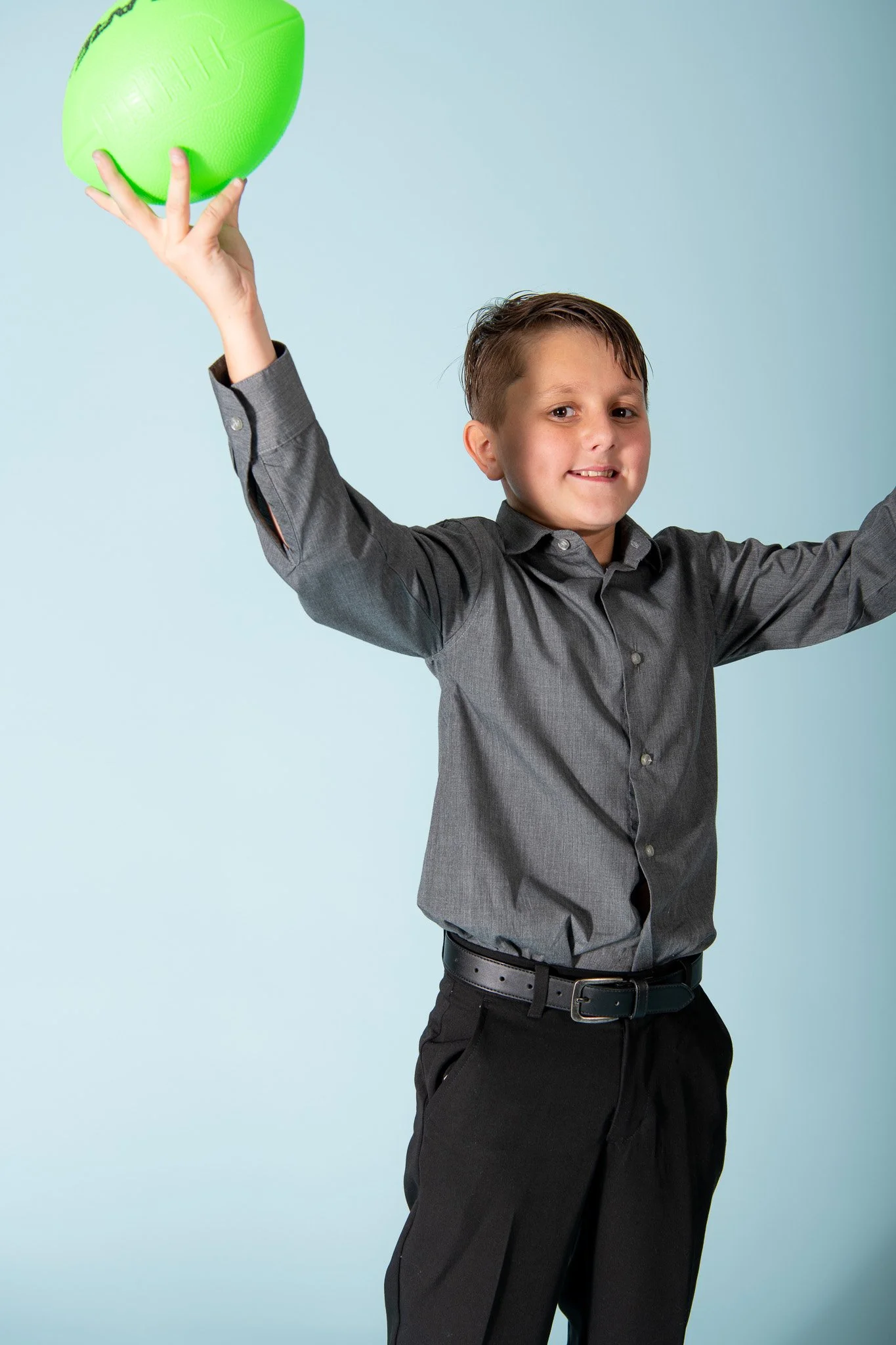 A young boy in a gray dress shirt and black pants holding a green football in the air, smiling, against a light blue background.