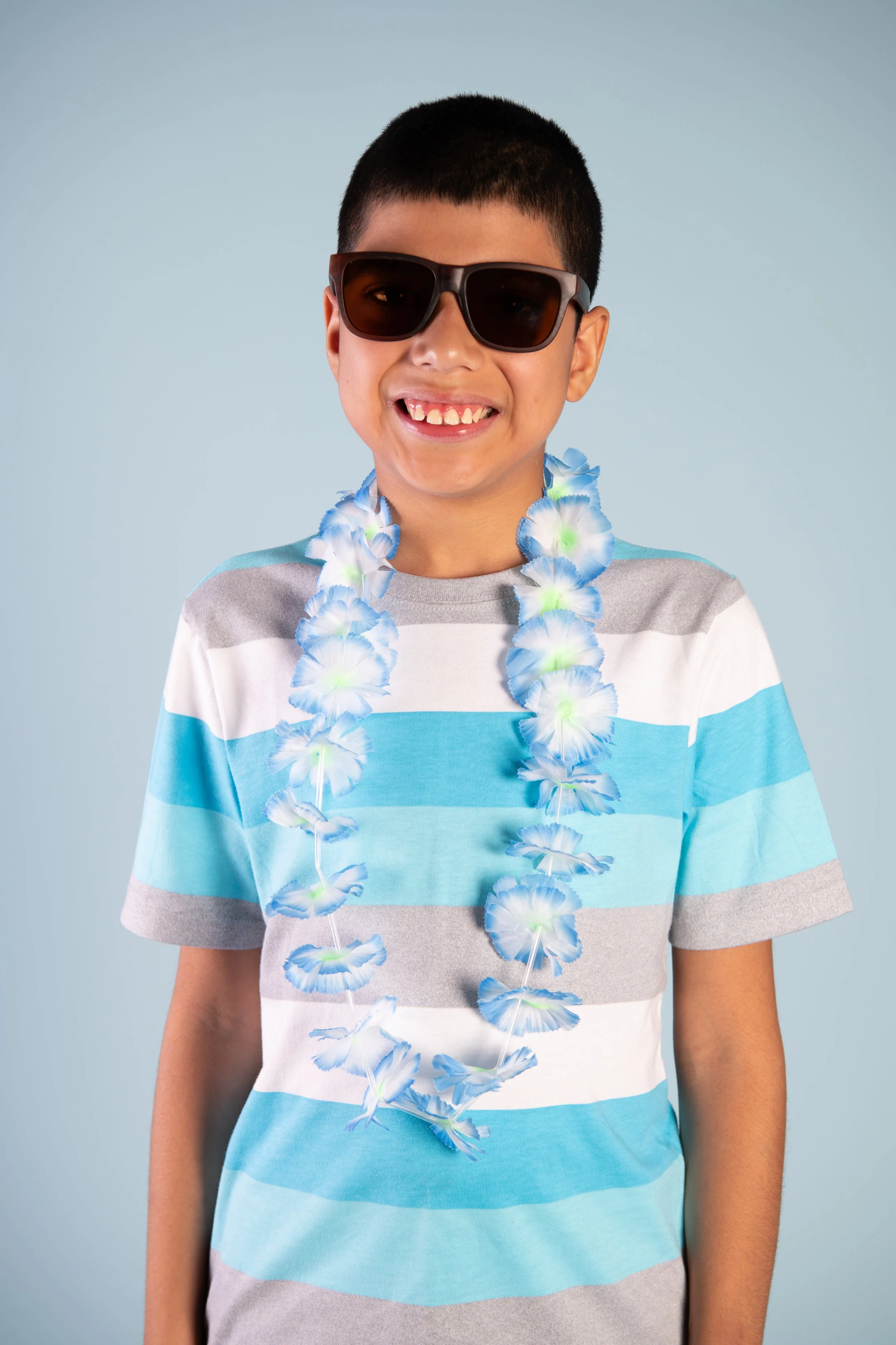 Young boy smiling, wearing sunglasses, a flower lei, and a striped shirt, against a light blue background.