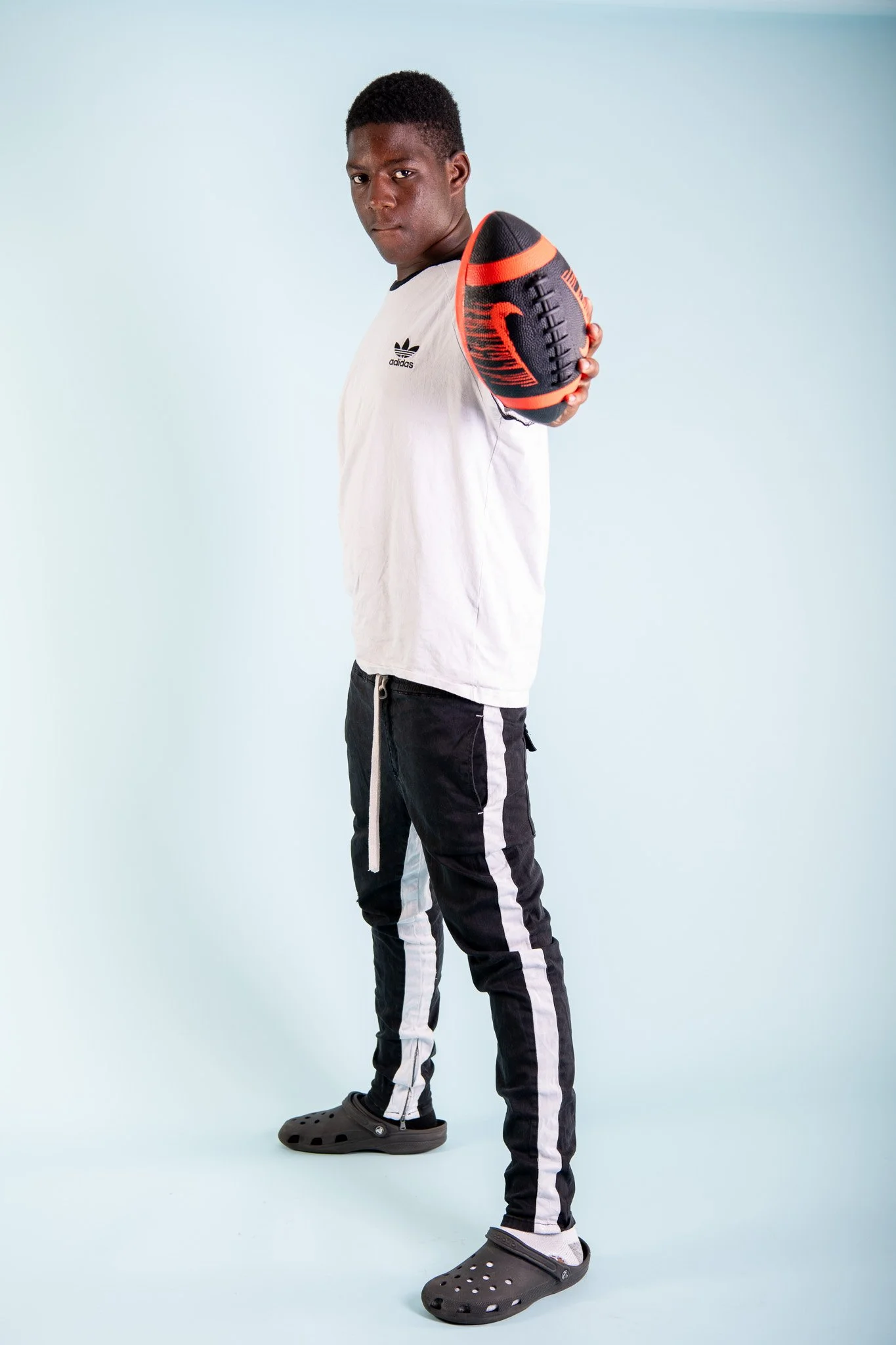 A young man with dark skin and short hair holding a football in his right hand, standing against a light blue background. He is wearing a white Adidas t-shirt, black and white track pants, and gray Crocs.
