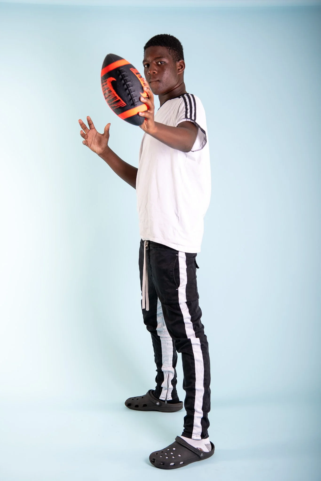 Young man holding a football, wearing casual clothes and Crocs, standing in front of a light blue background.