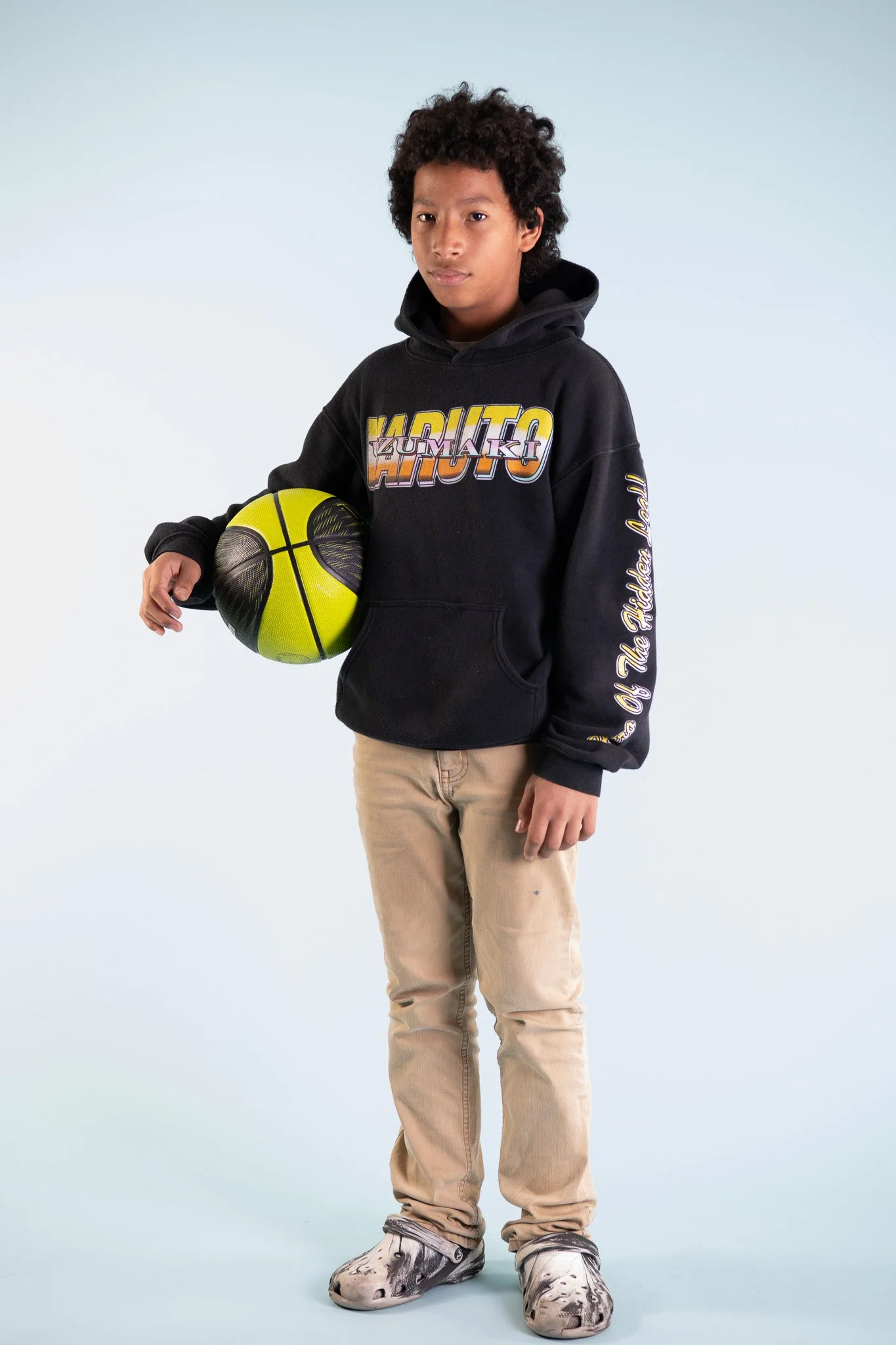 A young boy holding a yellow and black basketball, standing on a plain background, wearing a black hoodie with colorful text and beige pants.