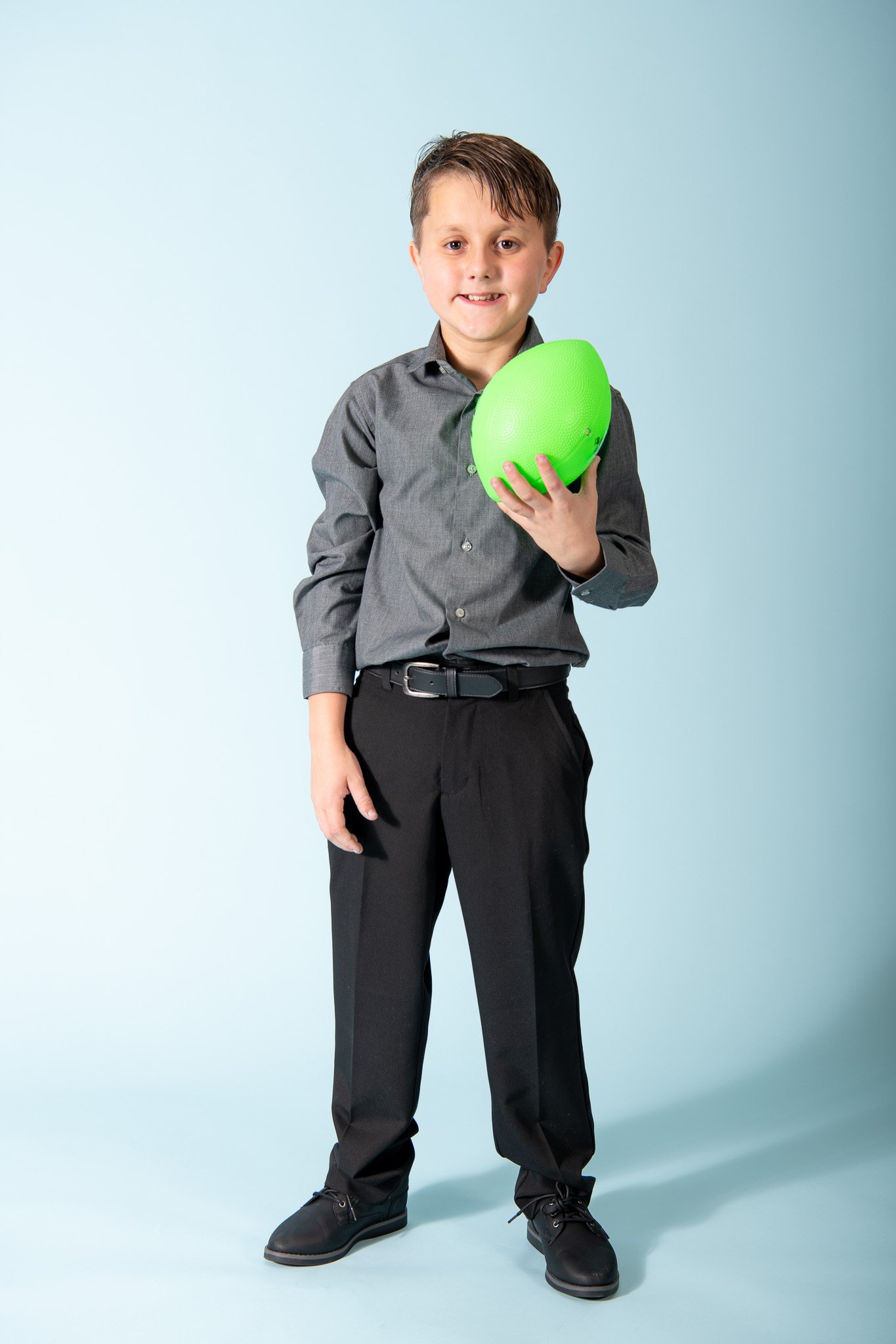 A young boy in a gray dress shirt and black pants holding a green football in his right hand, standing against a light blue background.