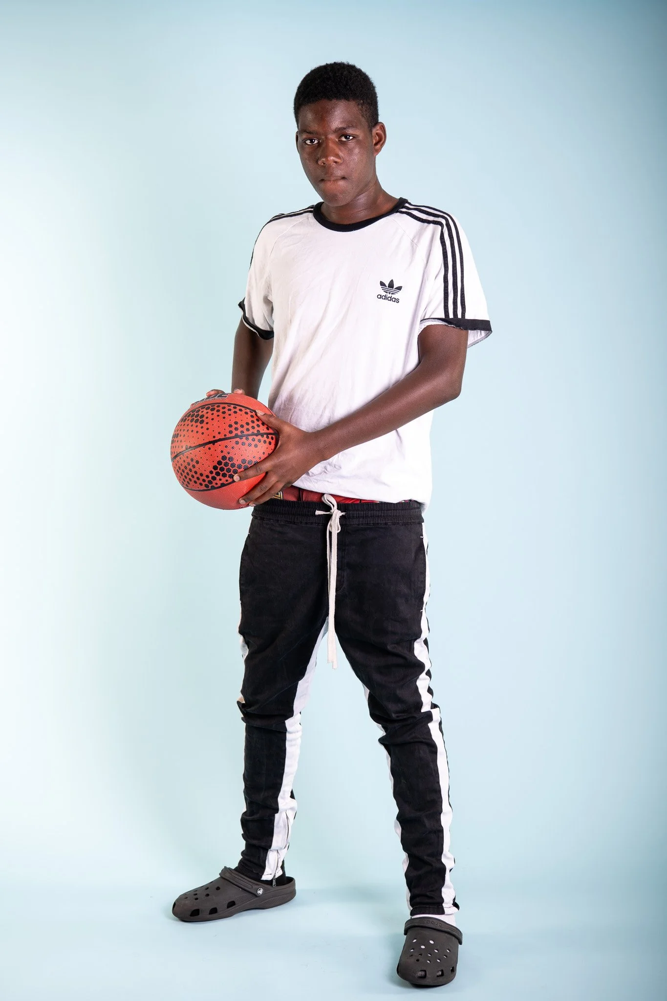 A teenage boy stands holding a basketball, dressed in casual sportswear, with a serious expression, against a light blue background.