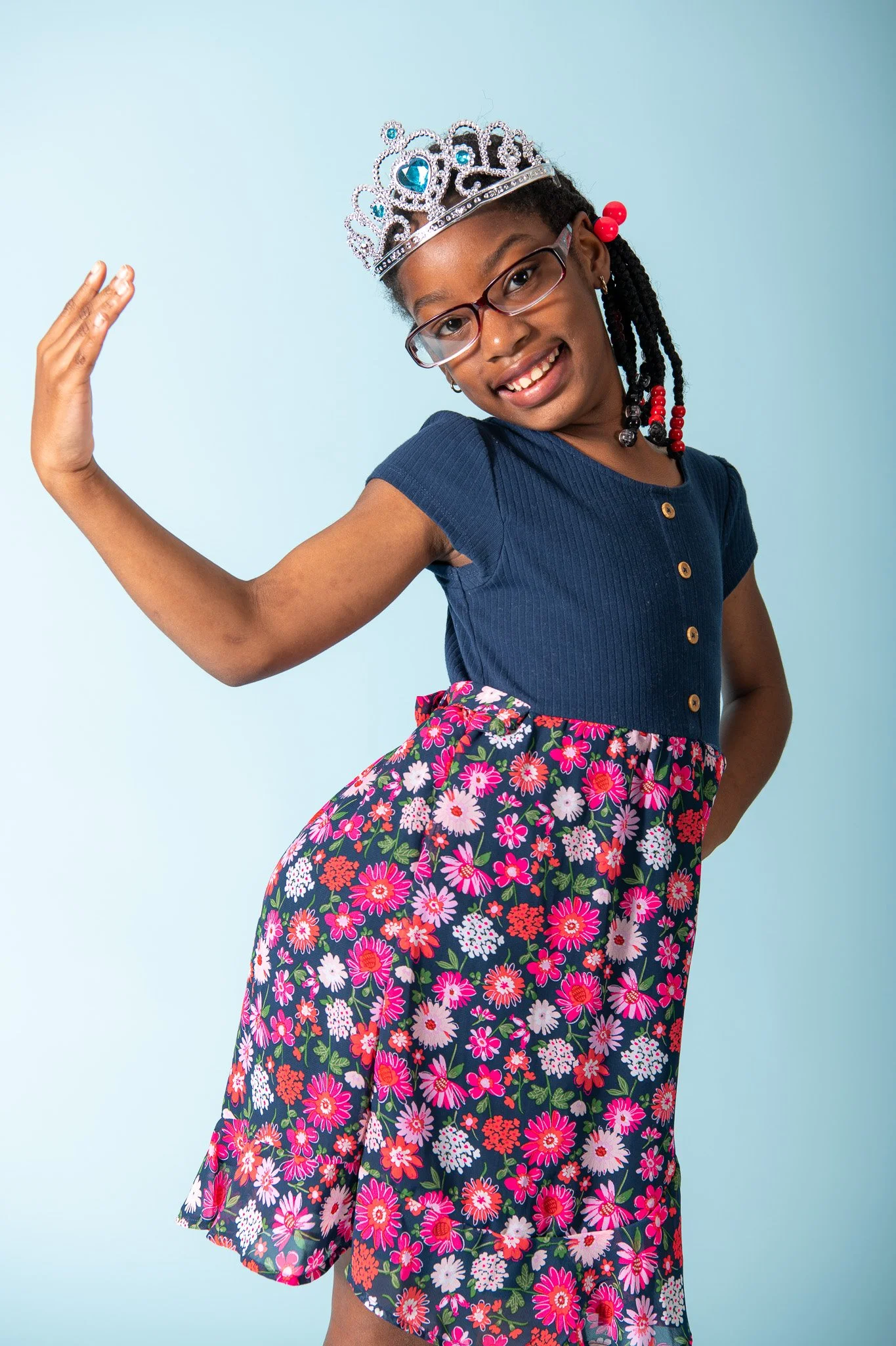 A young girl wearing glasses, a princess crown with blue gemstones, a navy blue shirt, and a floral skirt, posing with her head tilted, smiling, and raising her right arm against a light blue background.
