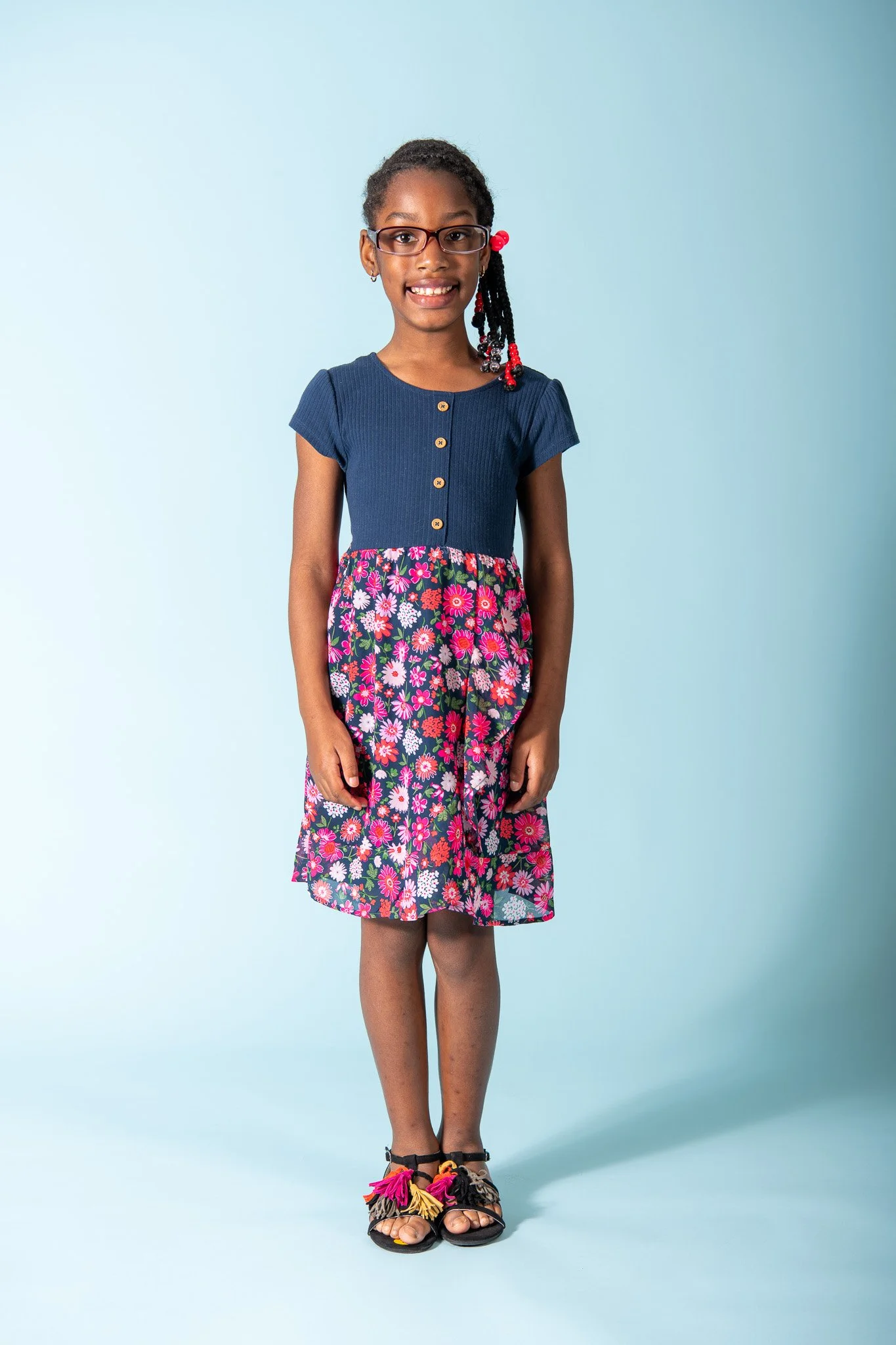 A young girl with glasses and braided hair, wearing a navy blue top with buttons and a colorful floral skirt, standing against a light blue background.