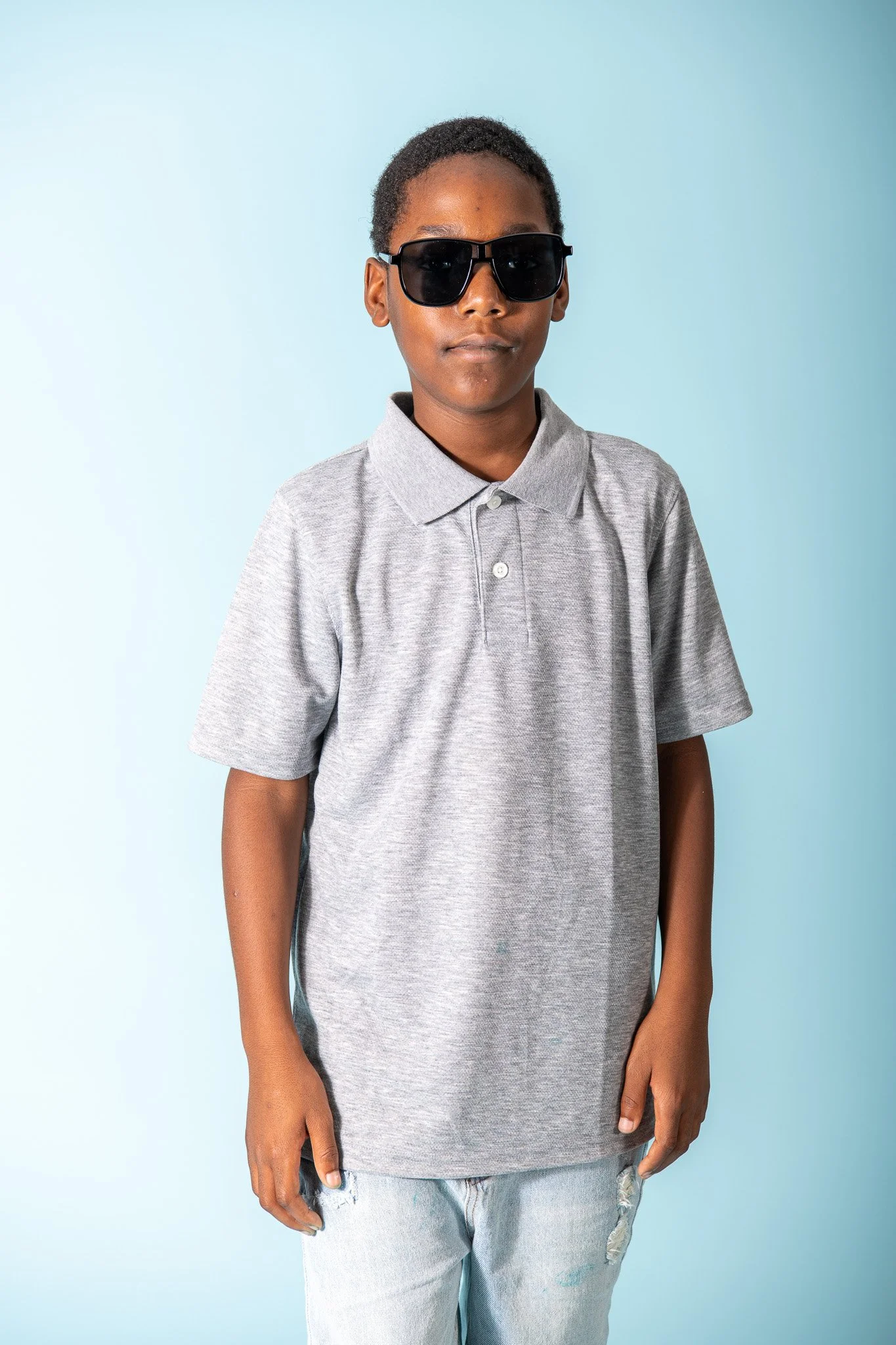 A young person with short hair wearing large black sunglasses, a gray polo shirt, and light blue ripped jeans standing against a light blue background.