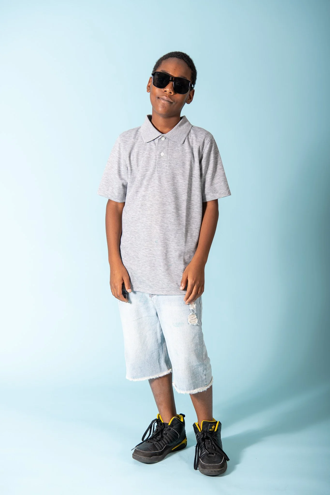 A young boy with dark sunglasses wearing a gray polo shirt, ripped denim shorts, and black sneakers with yellow accents standing against a light blue background.