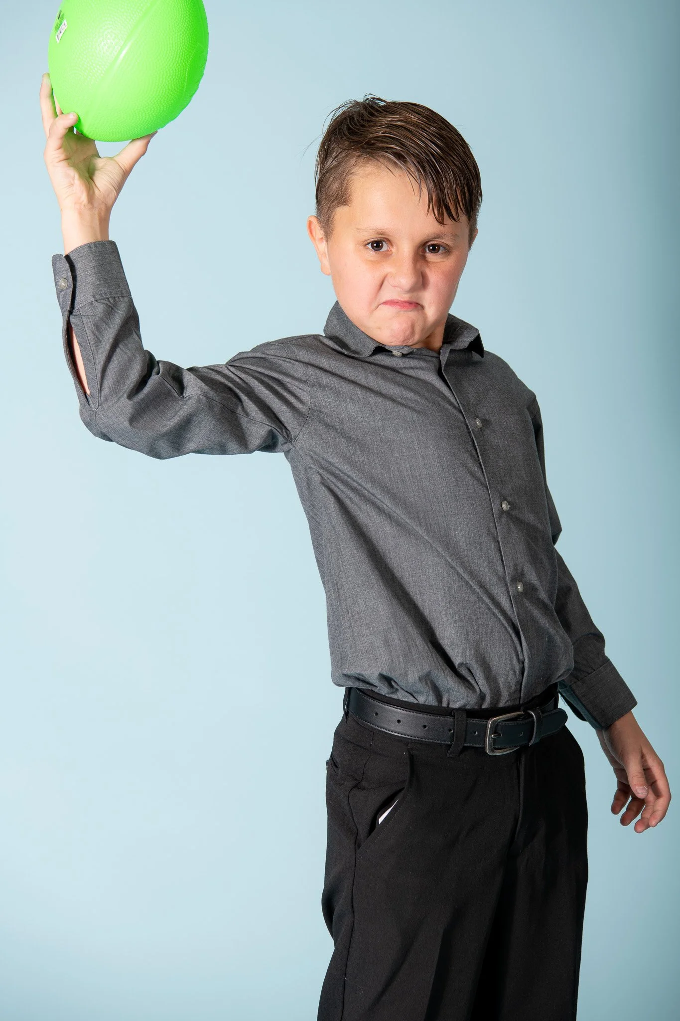 A young boy with a stern expression holding a green ball aloft, dressed in a gray button-up shirt and black pants against a light blue background.