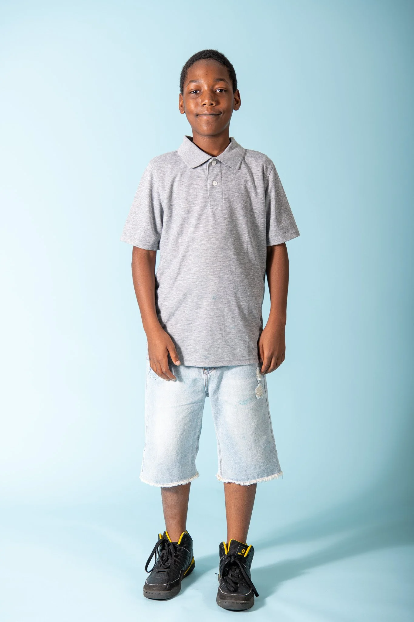 A young boy standing in front of a light blue background, wearing a gray polo shirt, distressed knee-length denim shorts, and black sneakers with yellow accents.