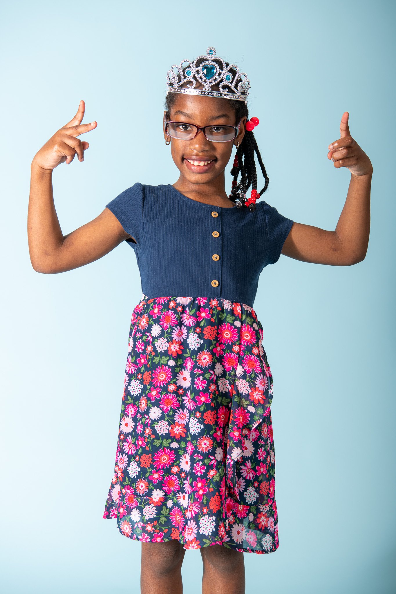 Young girl wearing glasses, a crown, a blue top, and a floral skirt, smiling and flexing her arms.