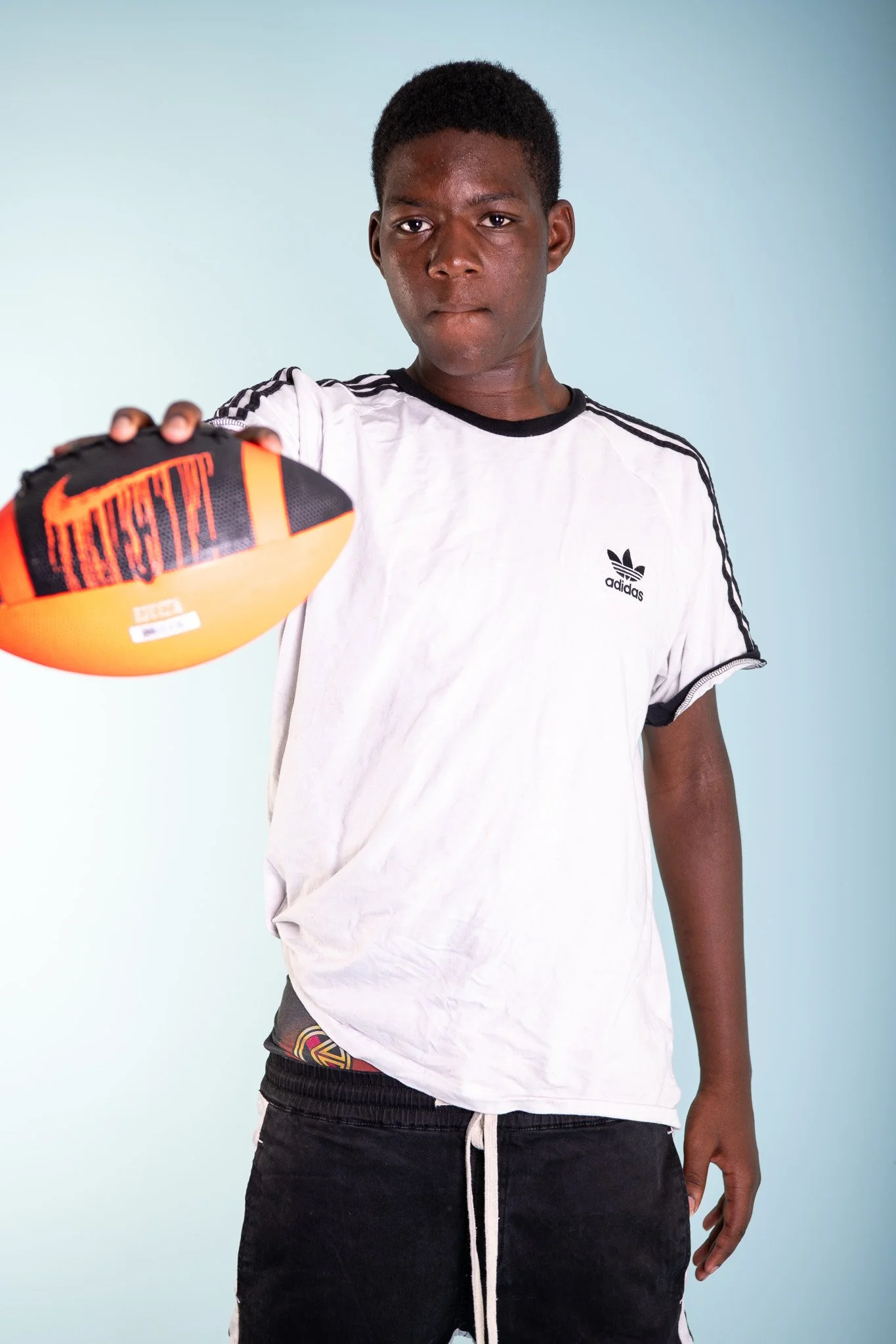A young man with dark skin and short black hair, wearing a white Adidas t-shirt, holding a Nike-branded football towards the camera, standing against a light blue background.