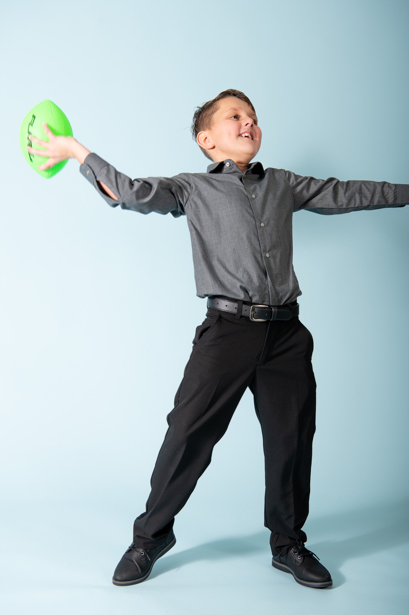 A young boy in a gray shirt and black pants holding a green football, smiling and stretching his arms out wide against a light blue background.
