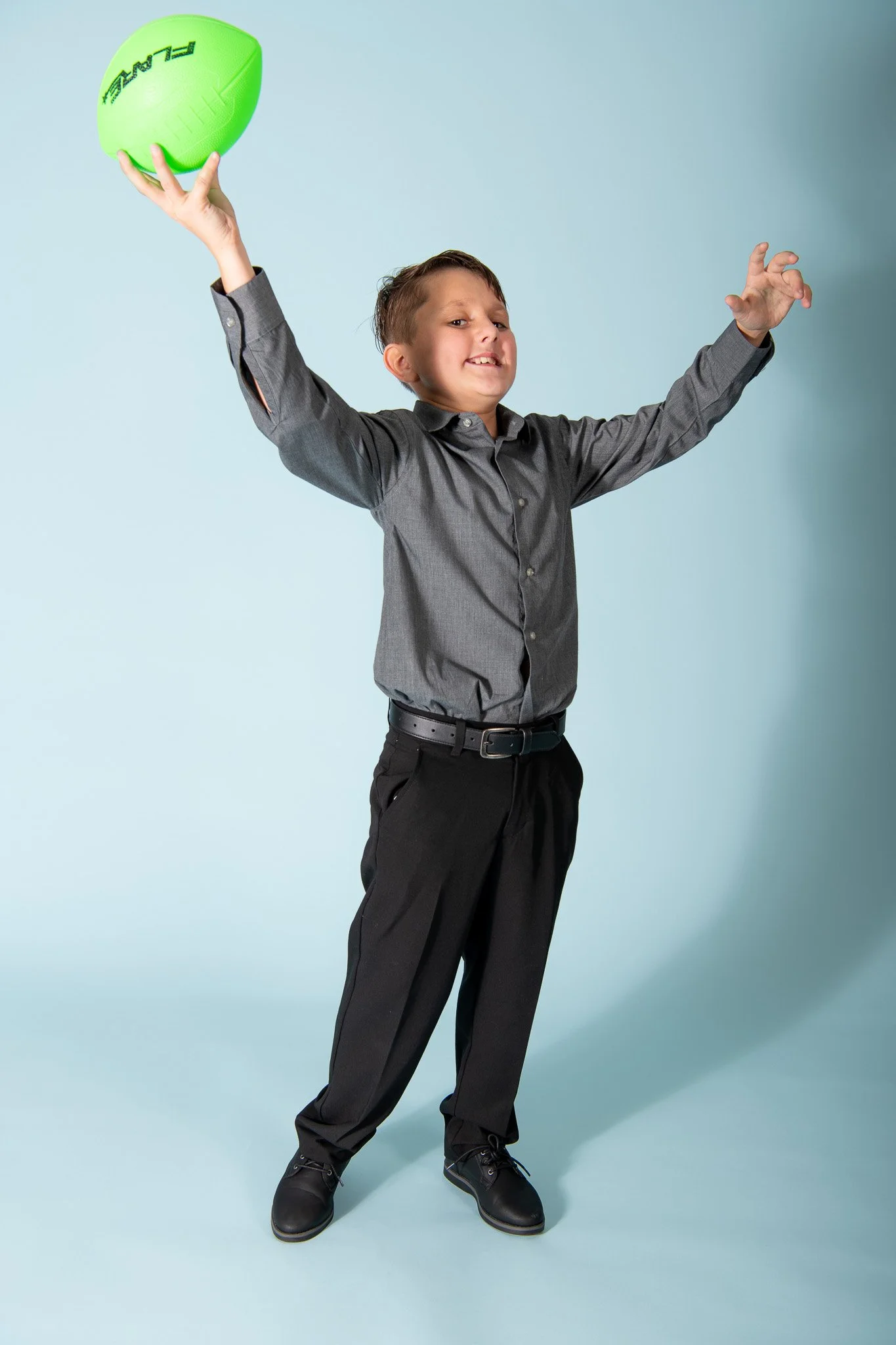 A young boy in a gray shirt and black pants holding a bright green football above his head, smiling and posing against a light blue background.