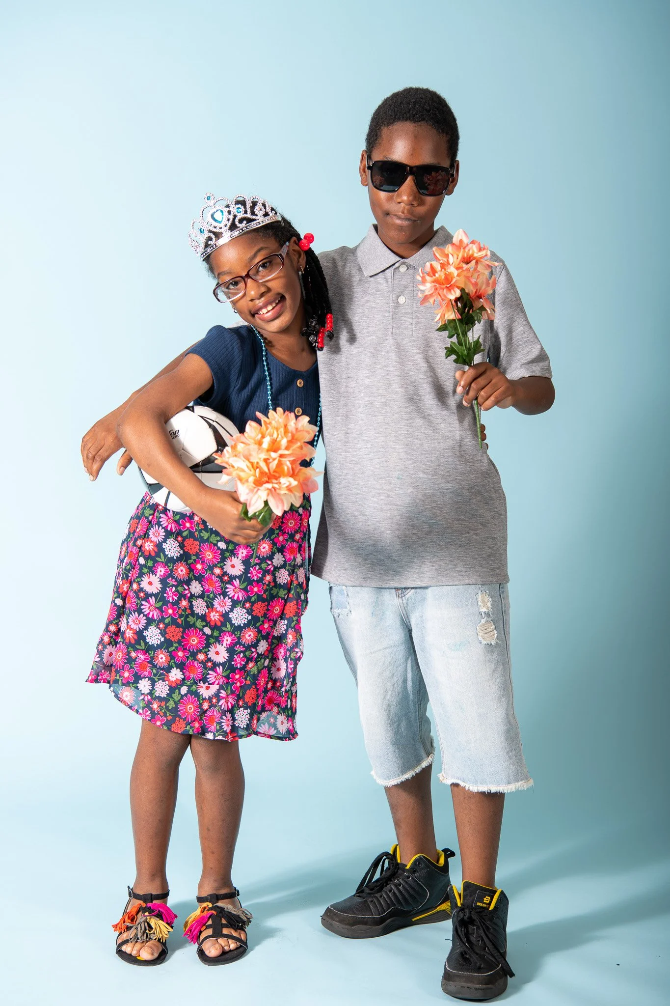 Two kids, a girl and a boy, standing together against a light blue background. The girl is smiling, wearing glasses, a tiara, a navy blue top, a floral skirt, and colorful sandals, holding a soccer ball and a flower. The boy is wearing sunglasses, a 