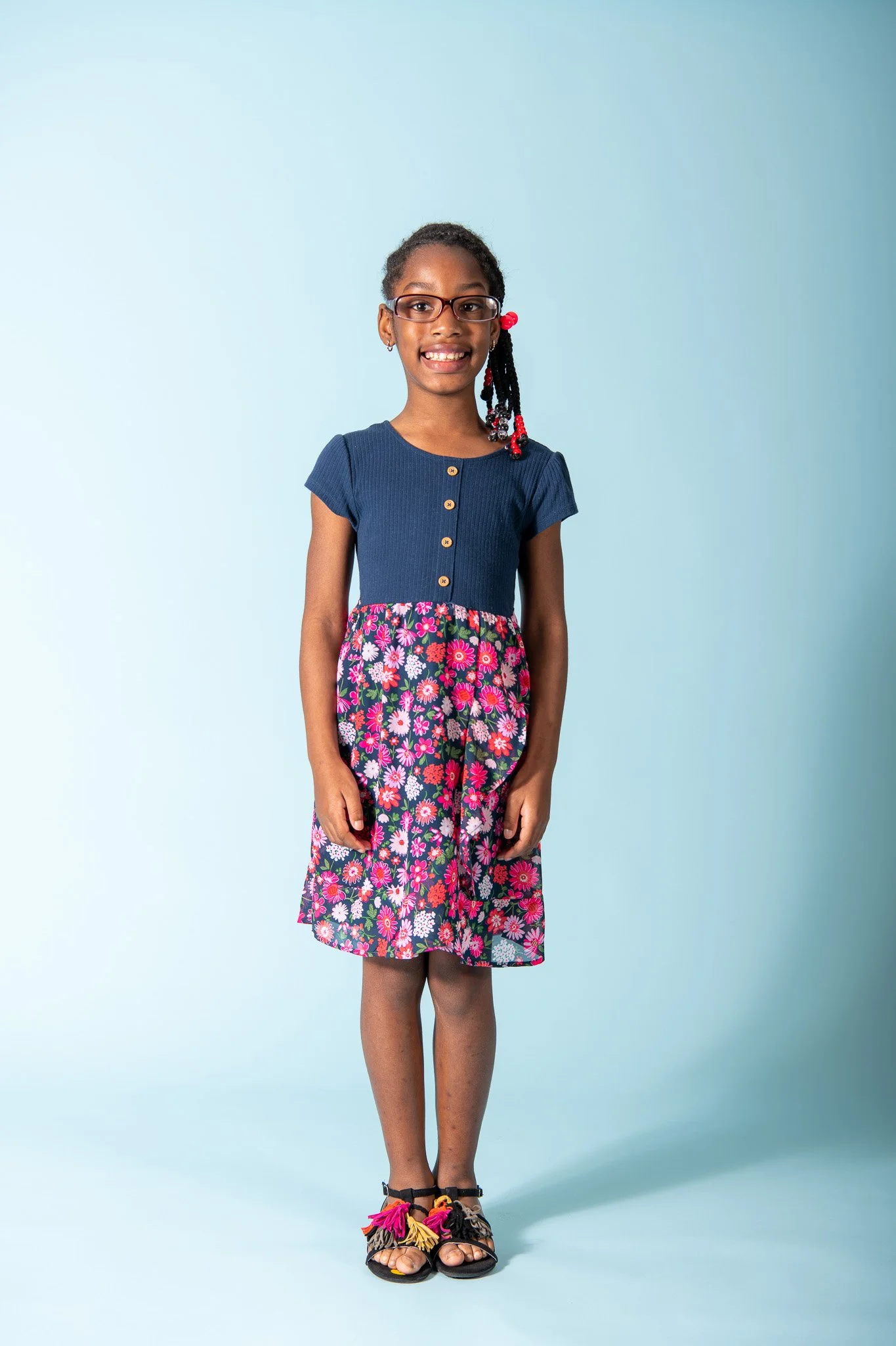 A young girl with glasses, wearing a navy blue top and a floral skirt, standing against a light blue background.