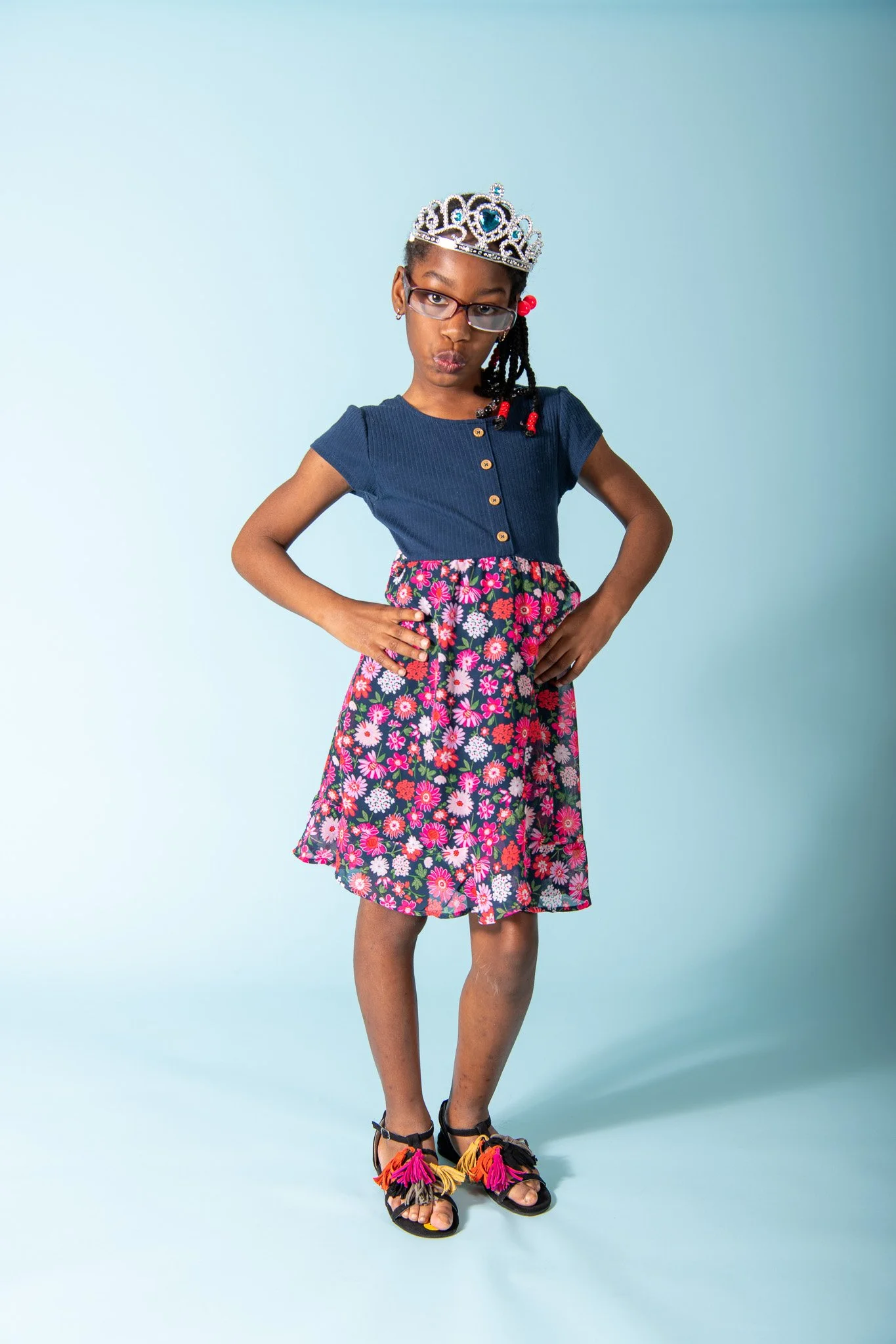 Young girl wearing glasses, a tiara, a blue top, a floral skirt, and colorful sandals, standing with hands on hips against a light blue background.