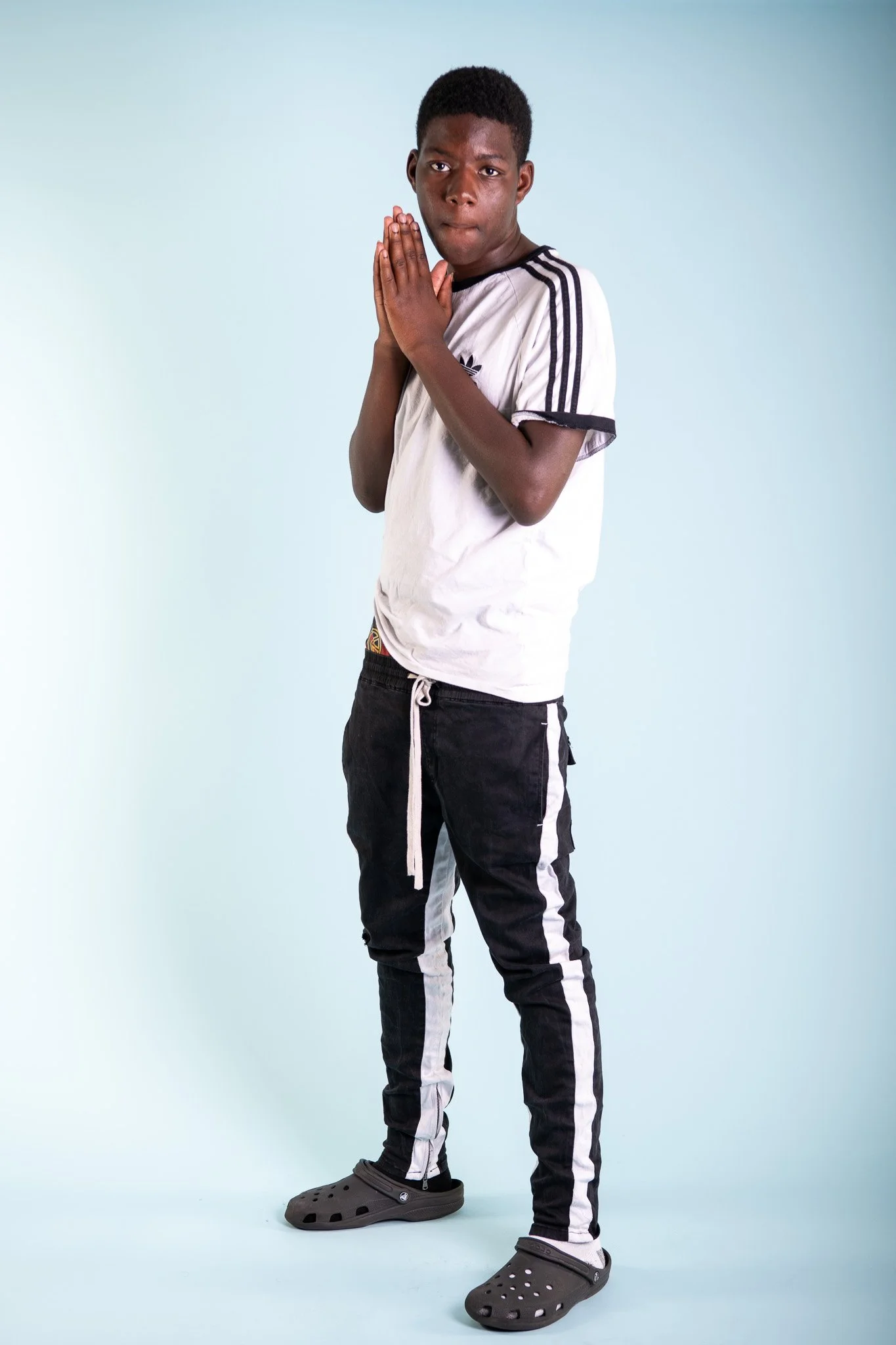 A young boy standing with his hands pressed together near his face, looking serious, wearing a white T-shirt with black stripes on the sleeves, black pants with white stripes, gray Crocs, and a white drawstring.