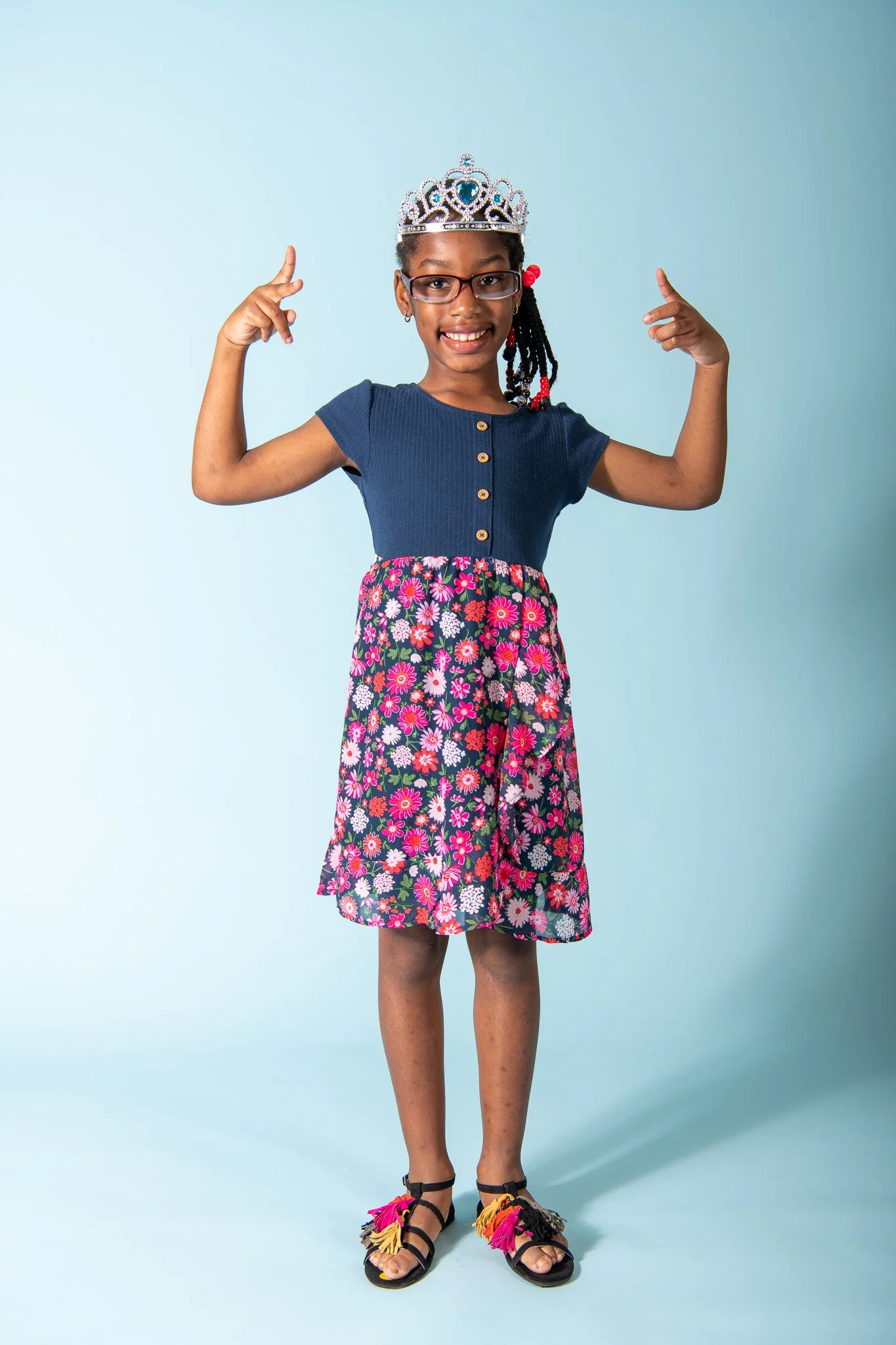 A young girl wearing glasses, a necklace, and a tiara, smiling and posing with her arms flexed, wearing a blue top, a floral skirt, and colorful sandals against a light blue background.