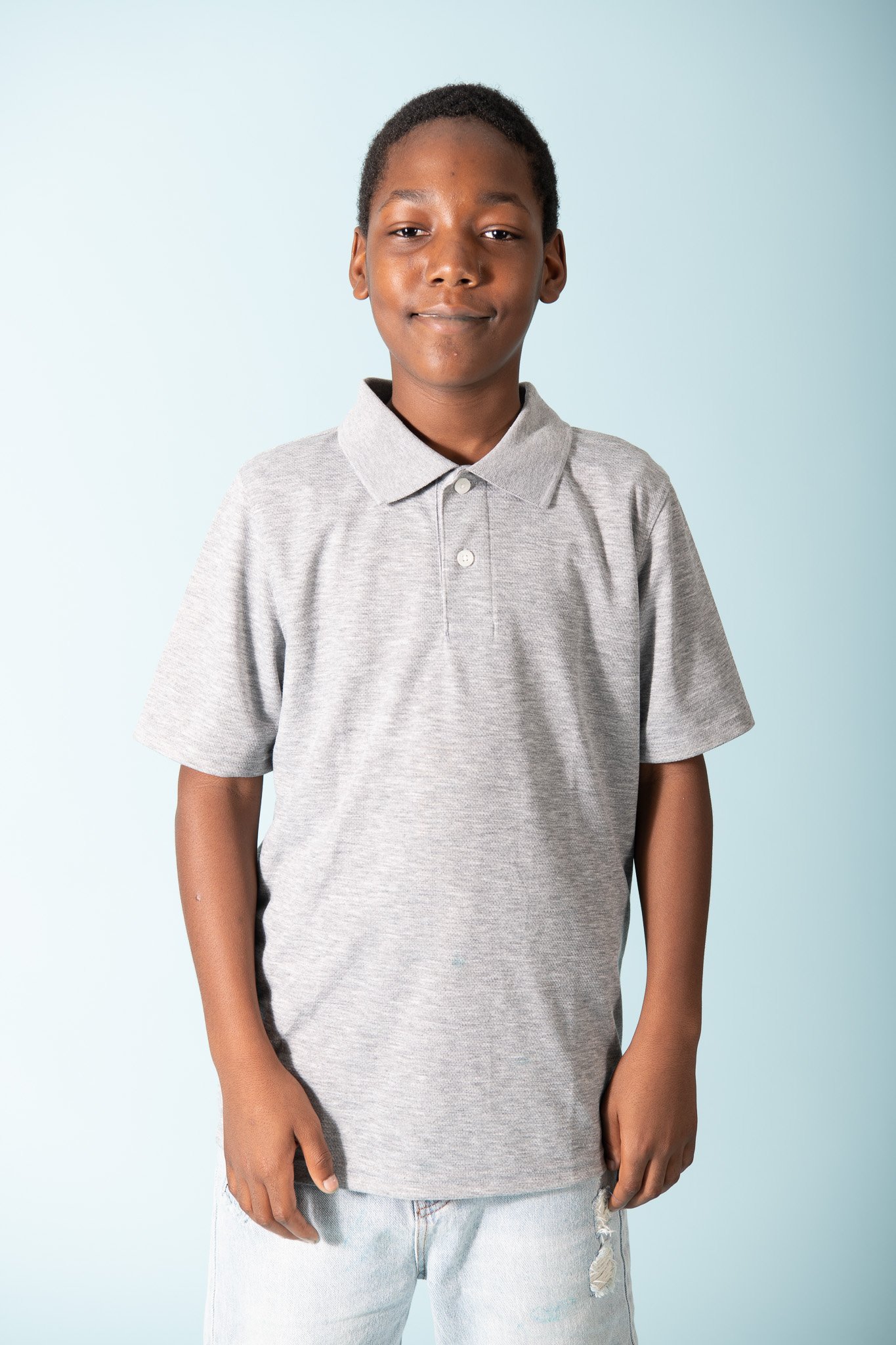 A young boy standing against a light blue background, wearing a gray polo shirt and light jeans.