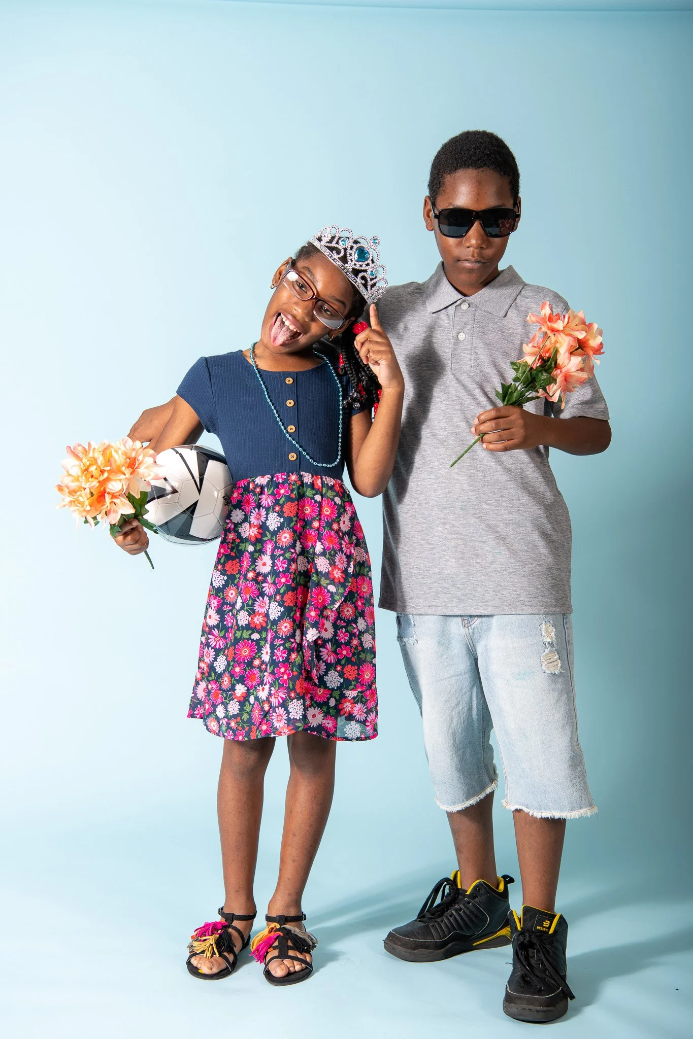 Two children, a girl and a boy, stand together against a light blue background. The girl is wearing a blue top, floral skirt, and sandals, holding a flower and a soccer ball, with a crown on her head and sticking out her tongue. The boy is wearing su