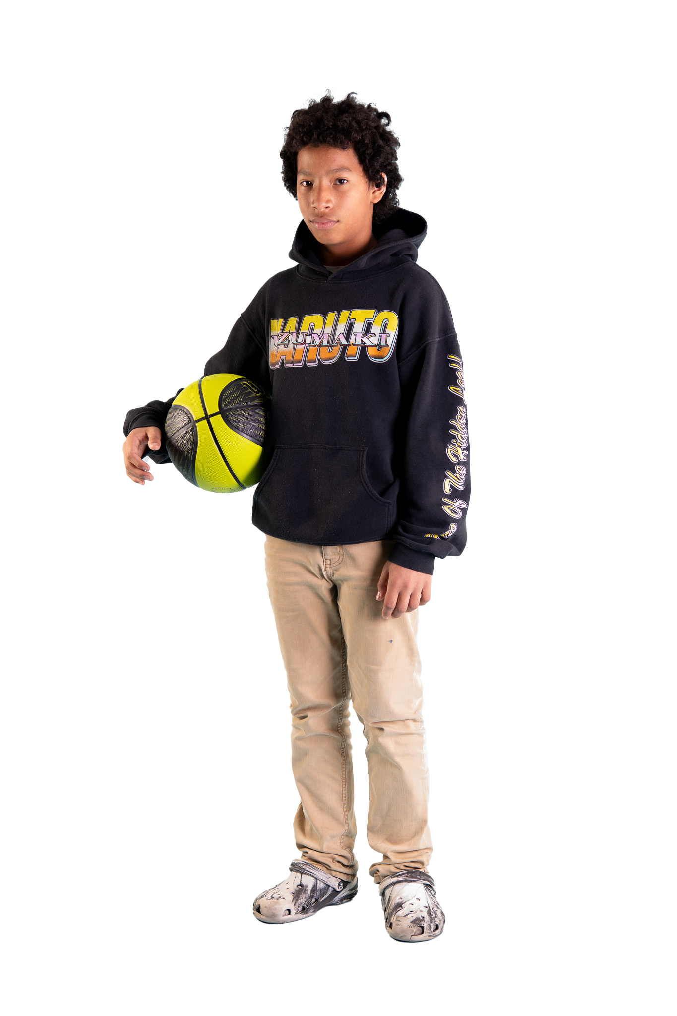A young boy holding a yellow and black basketball, wearing a black hoodie with colorful text and beige pants, standing against a plain background.