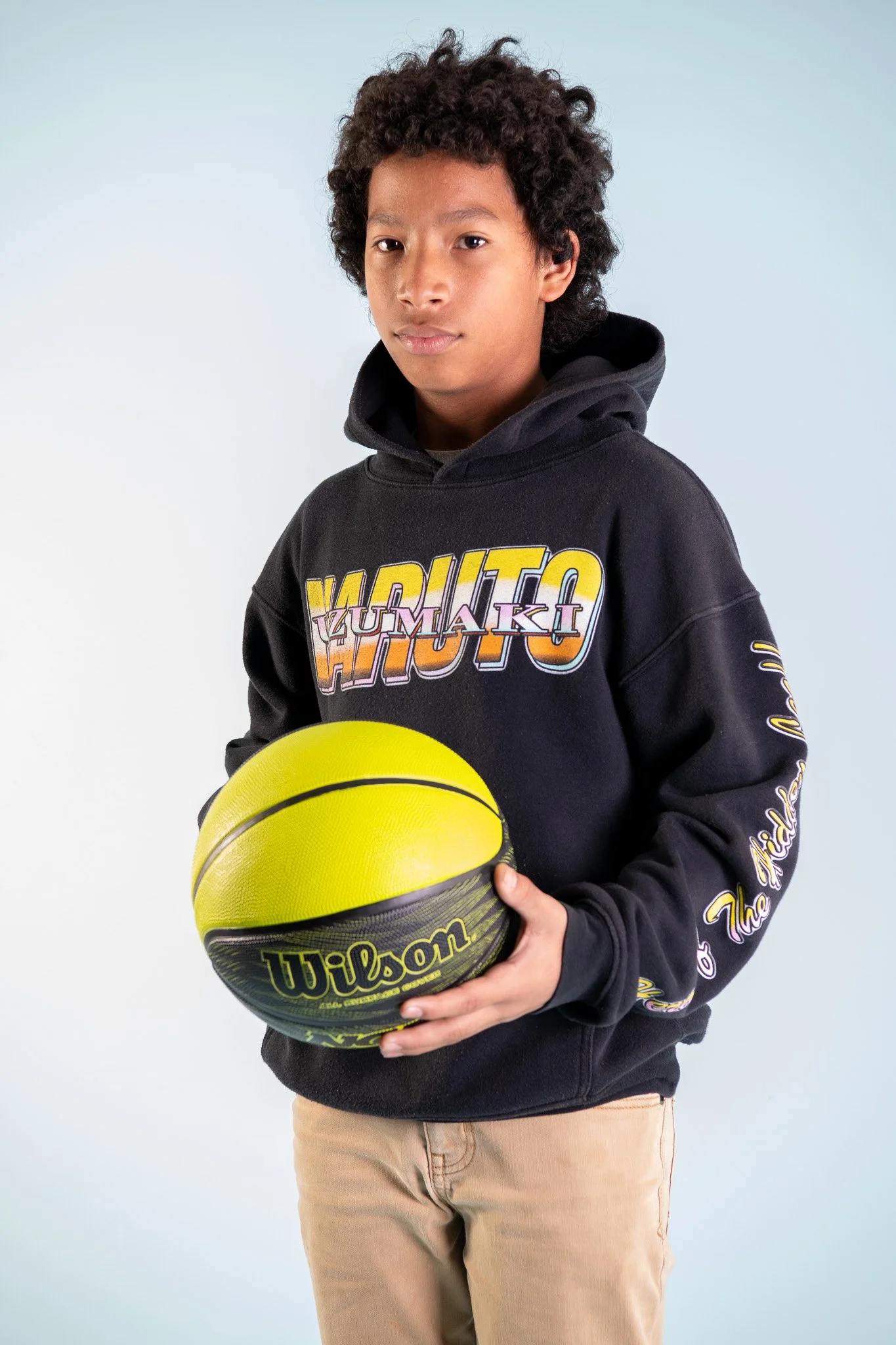 A young boy with curly hair holding a yellow and black Wilson basketball, wearing a black hoodie with colorful text and beige pants, standing against a plain light blue background.