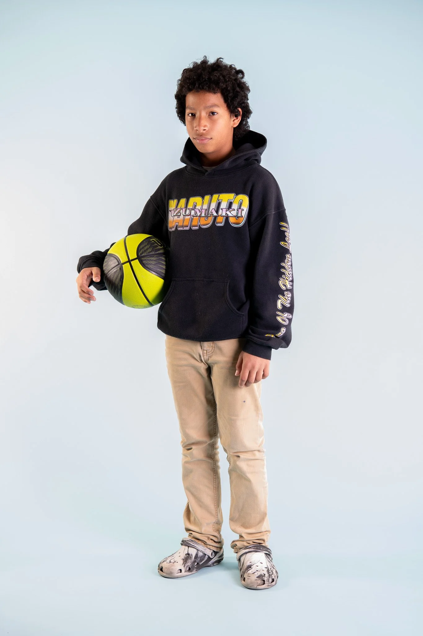 Teenage boy holding a yellow and black basketball, wearing a black hoodie with Naruto Kizumaki logo, beige pants, and patterned sneakers, standing against a plain light blue background.