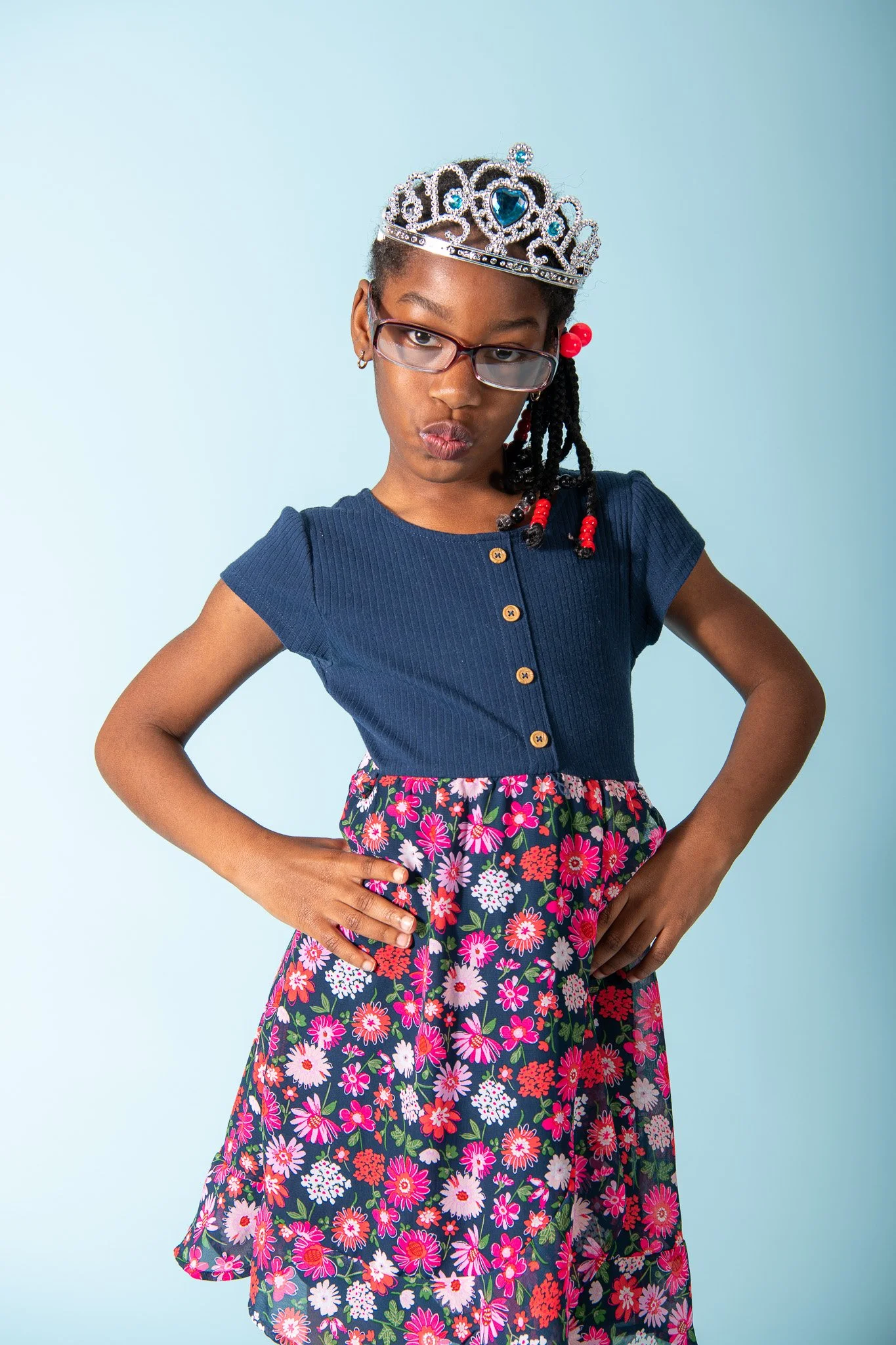 Young girl wearing glasses, a crown, a blue top, and a floral skirt, posing with hands on hips against a light blue background.