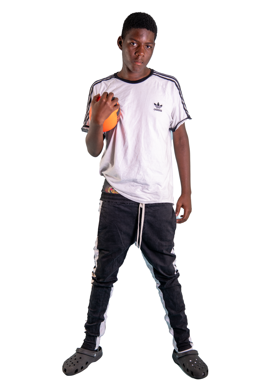 Young boy wearing a white Adidas t-shirt, black pants, and Crocs, holding an orange ball against his shoulder.