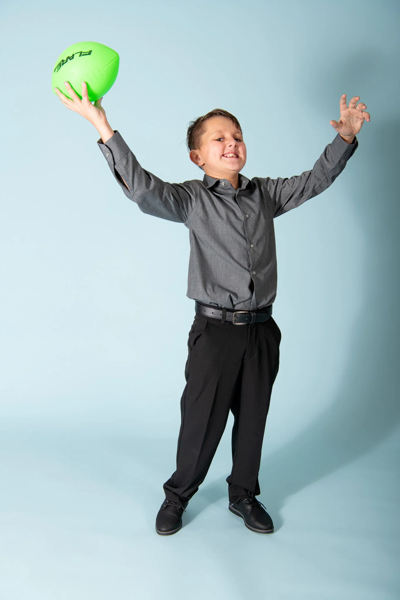 A young boy in a gray dress shirt and black pants holding a green football, posing with arms outstretched against a light blue background.