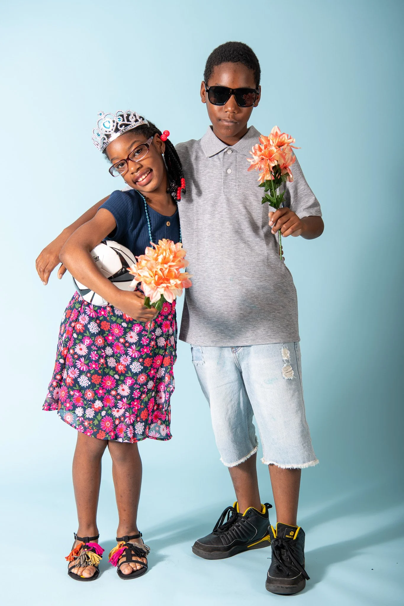 Two children standing together against a light blue background, smiling and holding flowers. The girl is wearing a tiara, glasses, a blue top, a floral skirt, and beaded jewelry. She is holding a soccer ball and a flower. The boy is wearing sunglasse