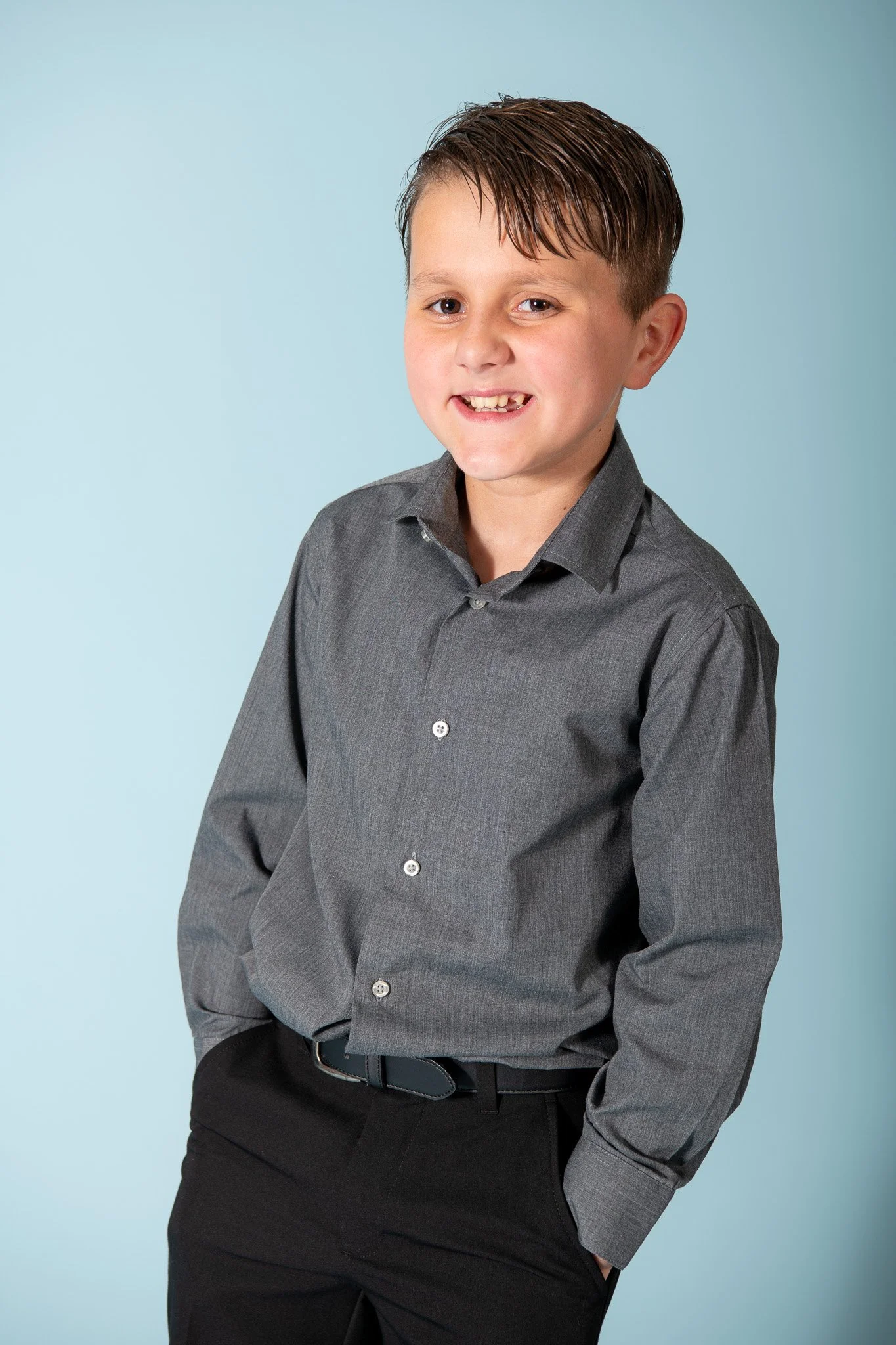 A young boy with a missing front tooth, wearing a gray button-down shirt and black pants, smiling with his hands in his pockets against a light blue background.