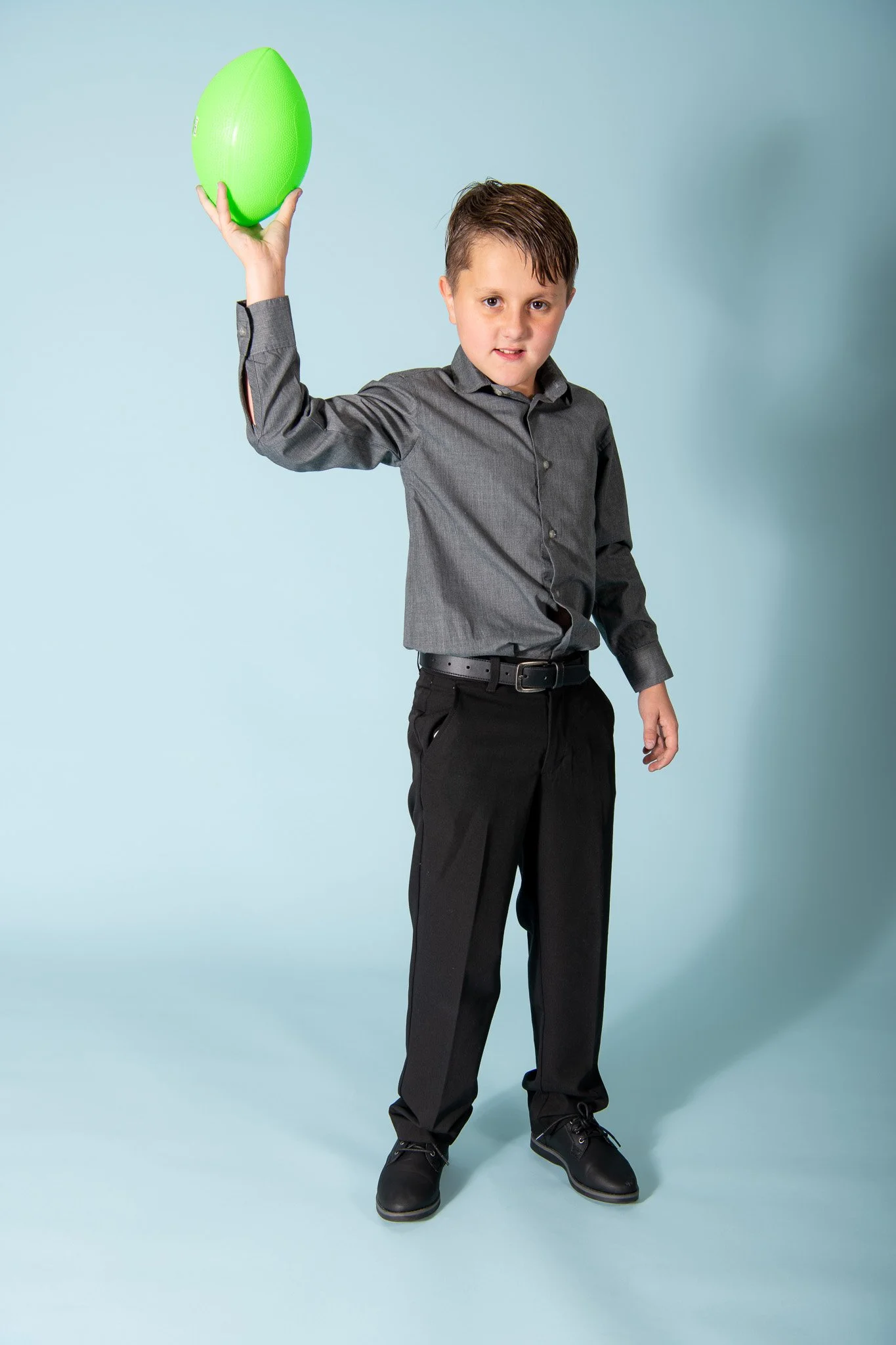 A young boy dressed in a gray dress shirt and black pants holding a green football in a studio with a light blue background.