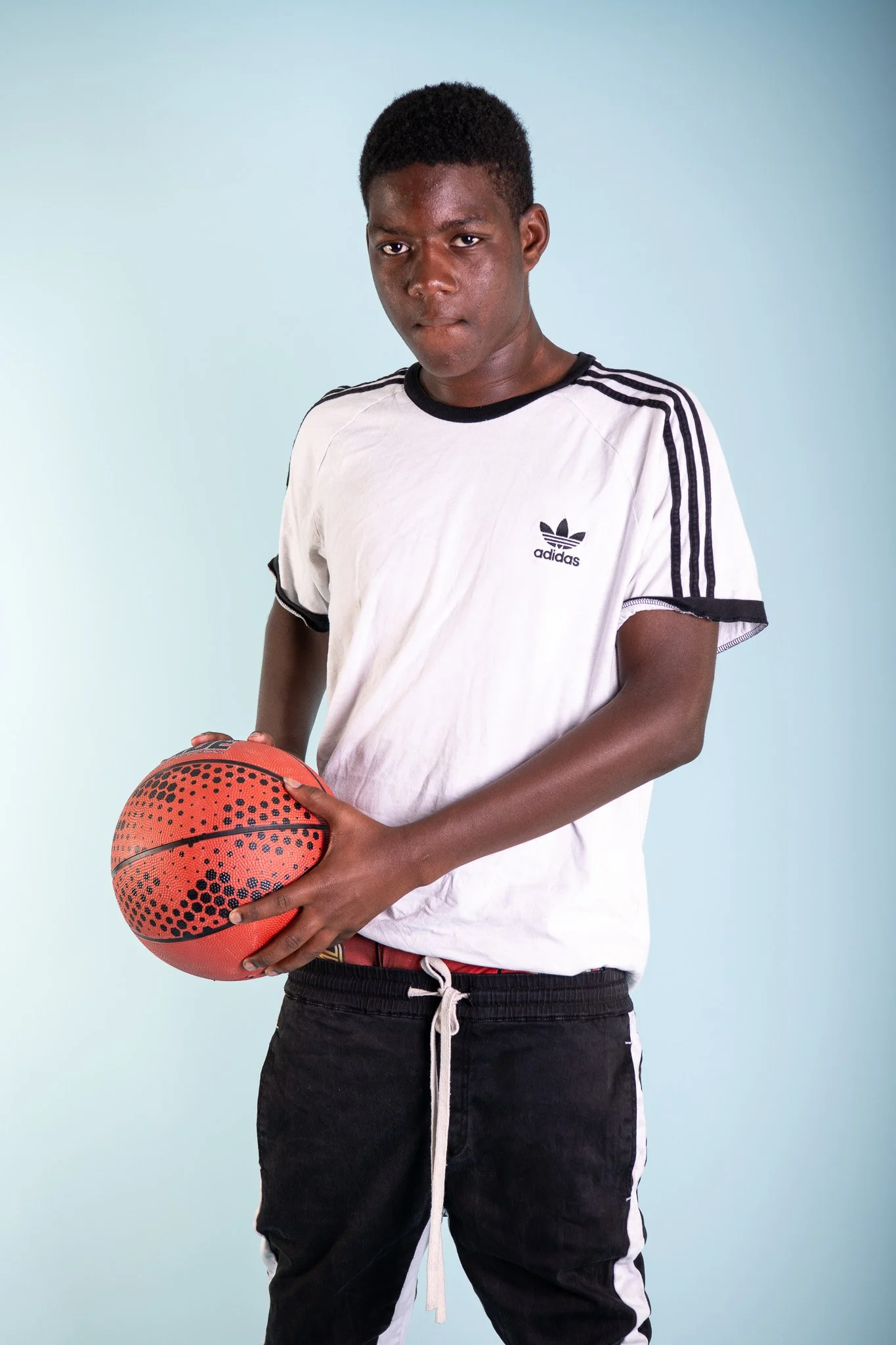 A young man standing with a basketball against a light blue background, wearing a white Adidas t-shirt and black pants.