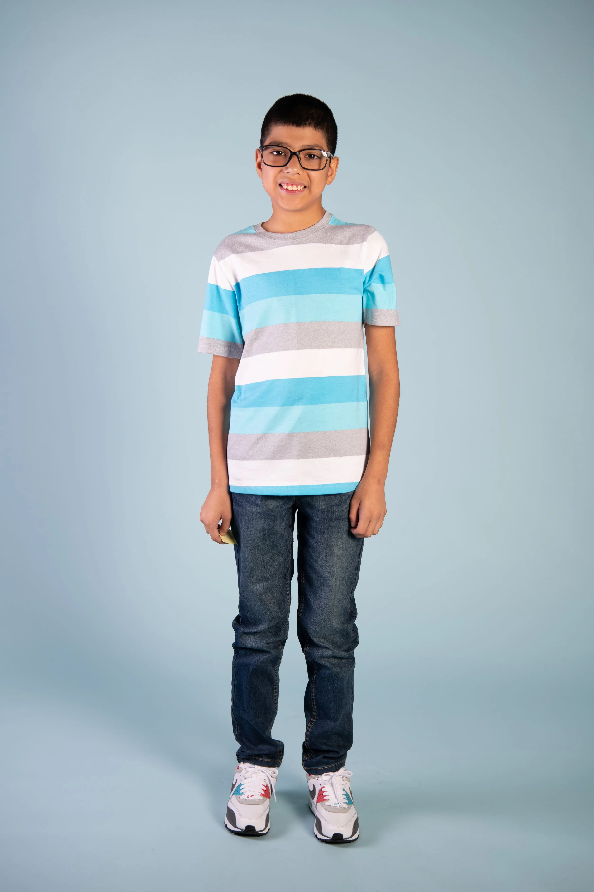 A young boy with short black hair wearing glasses, a striped blue, gray, and white T-shirt, dark blue jeans, and white sneakers with red, gray, and blue accents, standing against a light blue background.