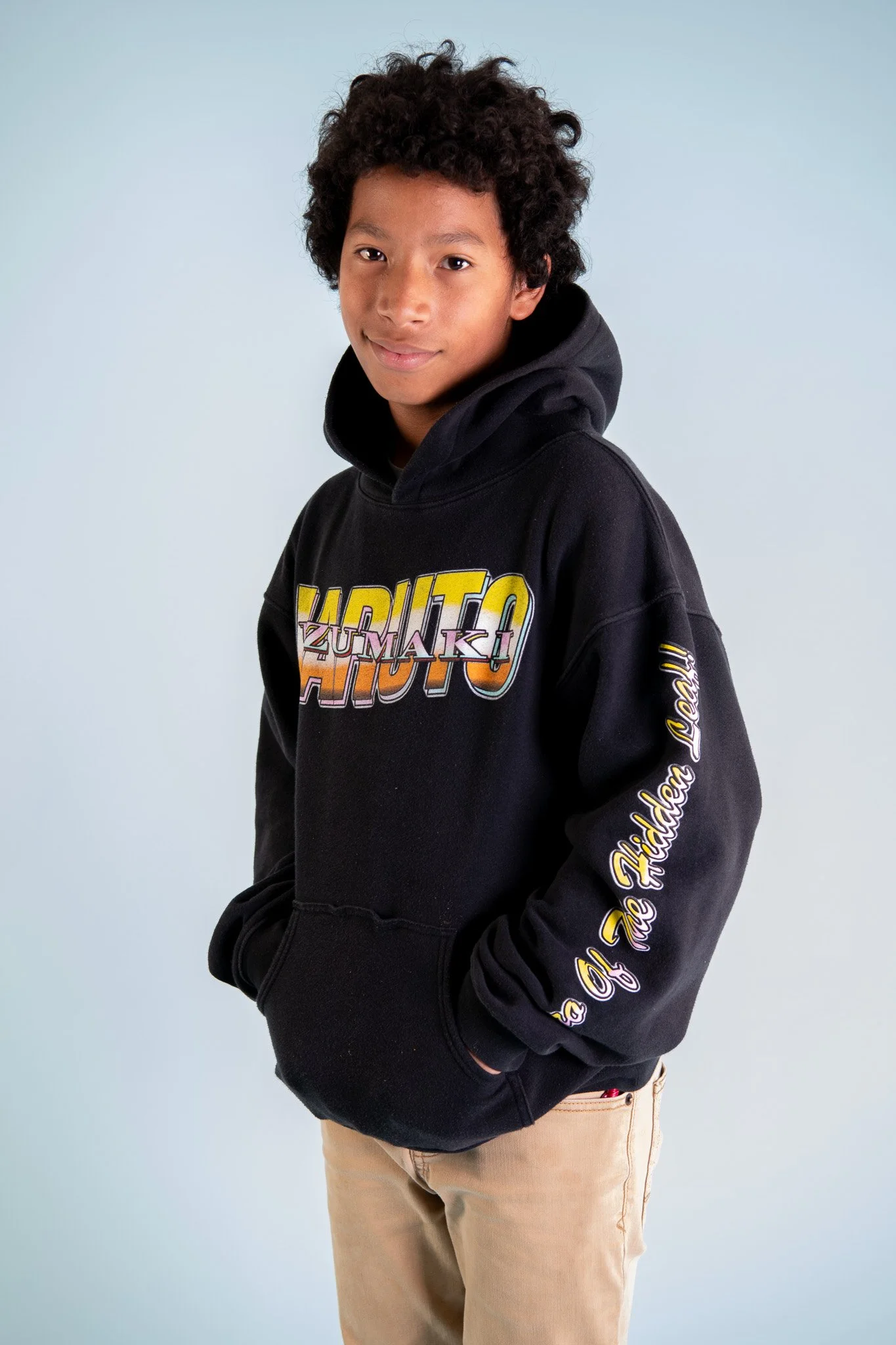 A teenage boy with curly hair wearing a black hoodie with colorful text and beige pants, standing against a light blue background.