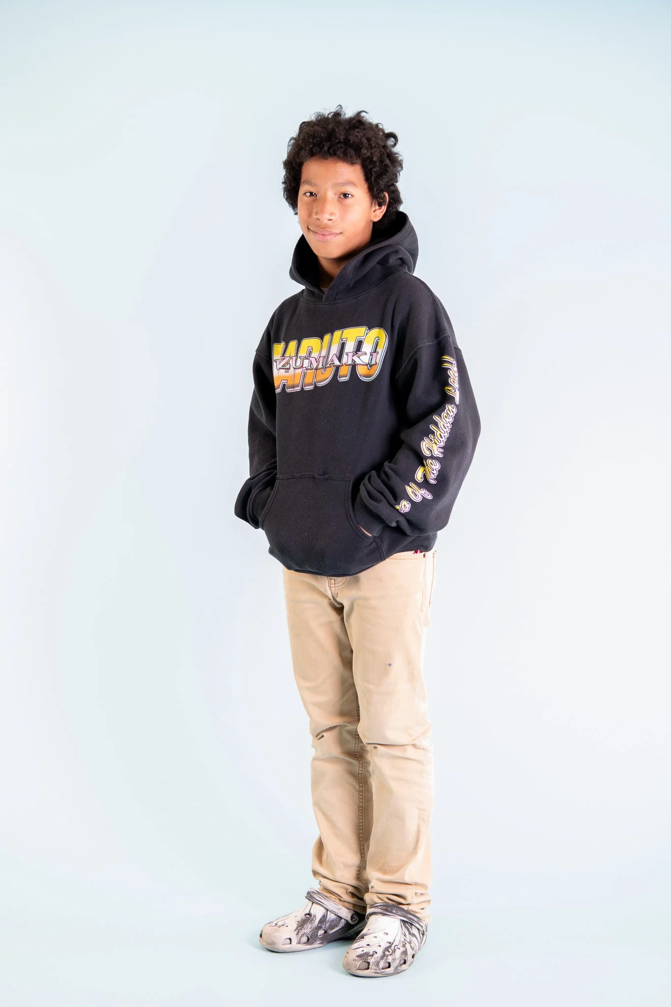 A young boy standing with hands in pockets, wearing a black hoodie with colorful graphic text, beige pants, and patterned shoes, against a plain white background.