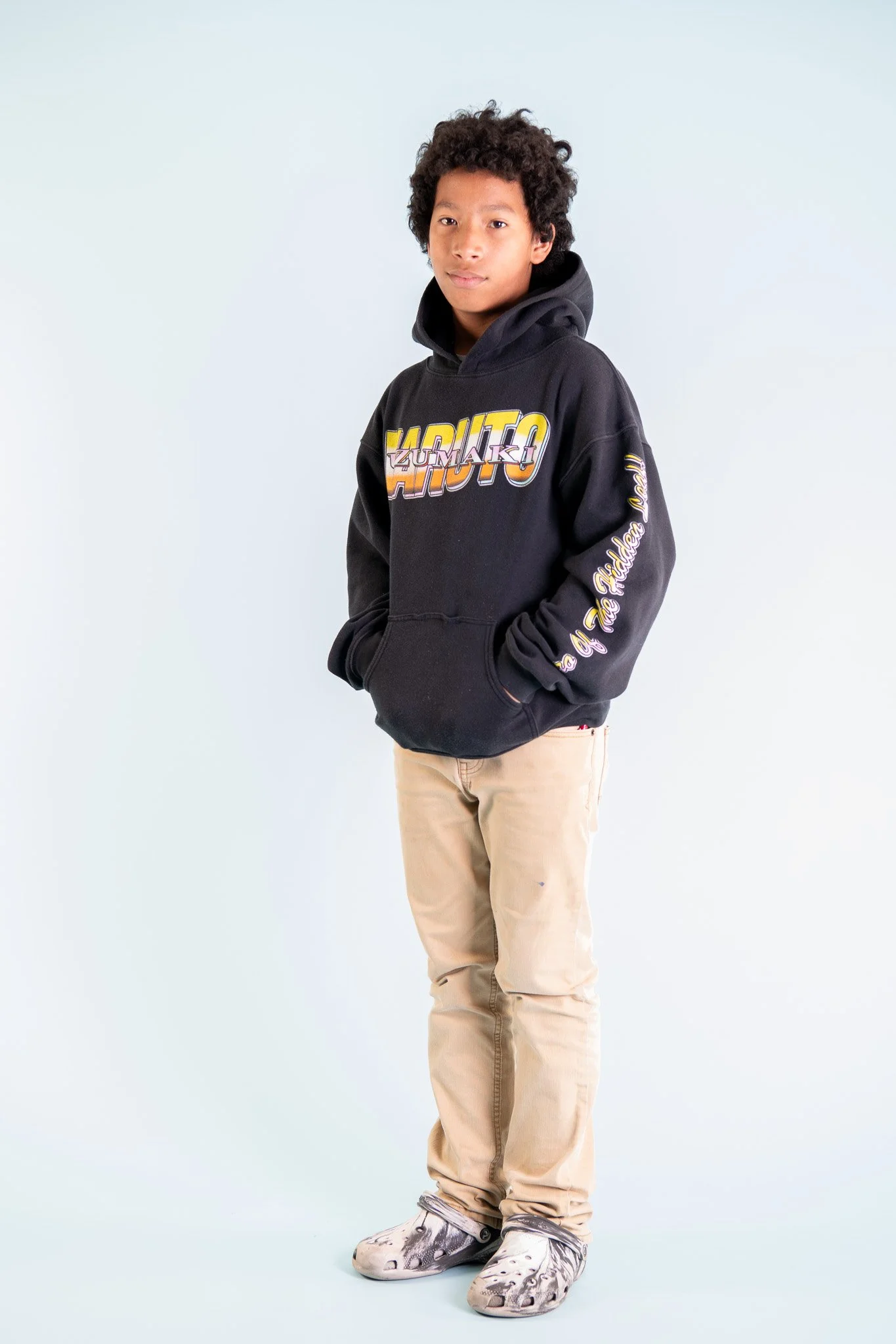 A young person with curly hair wearing a black hoodie with colorful text, tan pants, and unique footwear, standing against a plain light background.