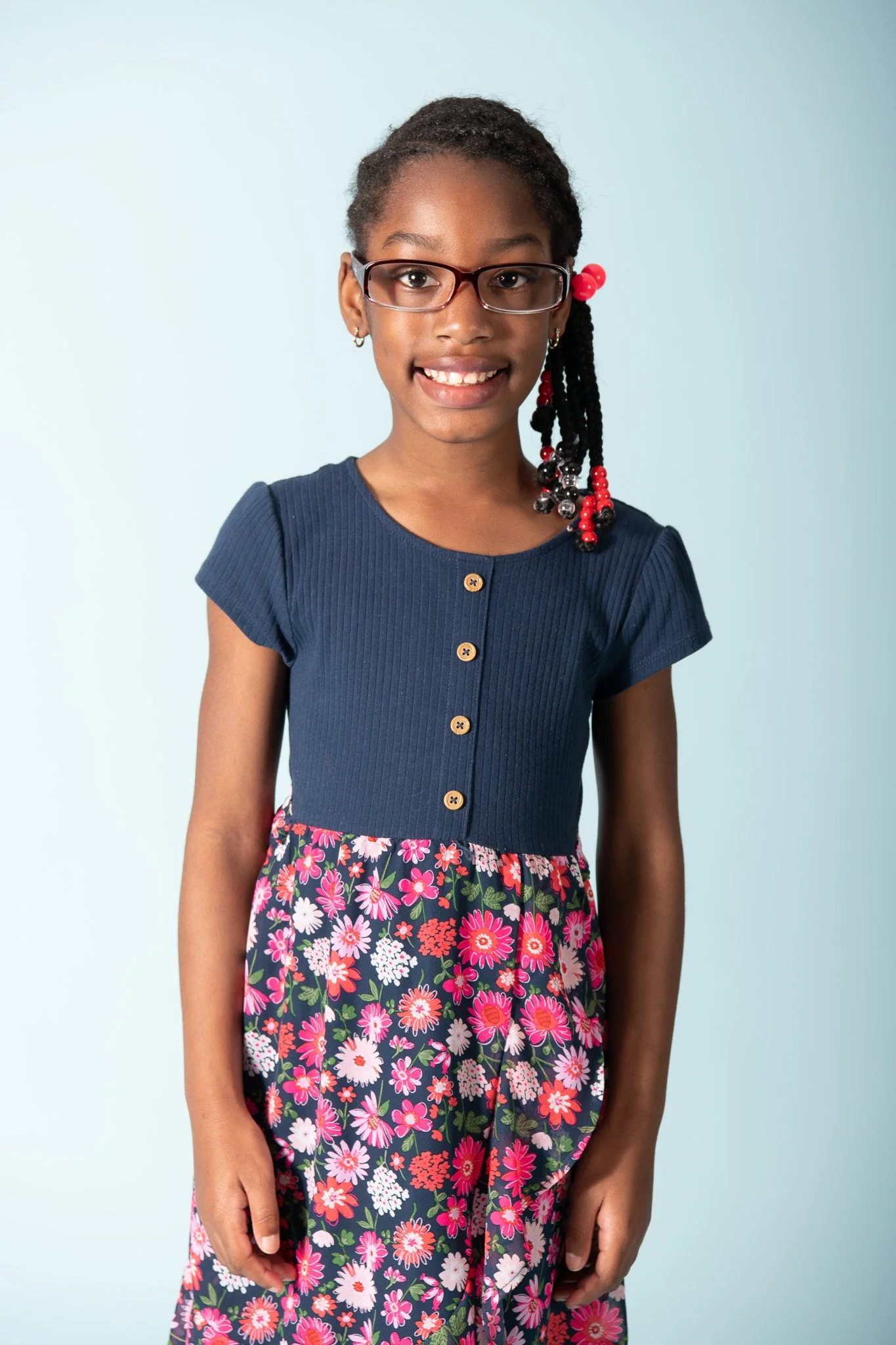 A young girl with glasses and a colorful dress, smiling at the camera against a light blue background.