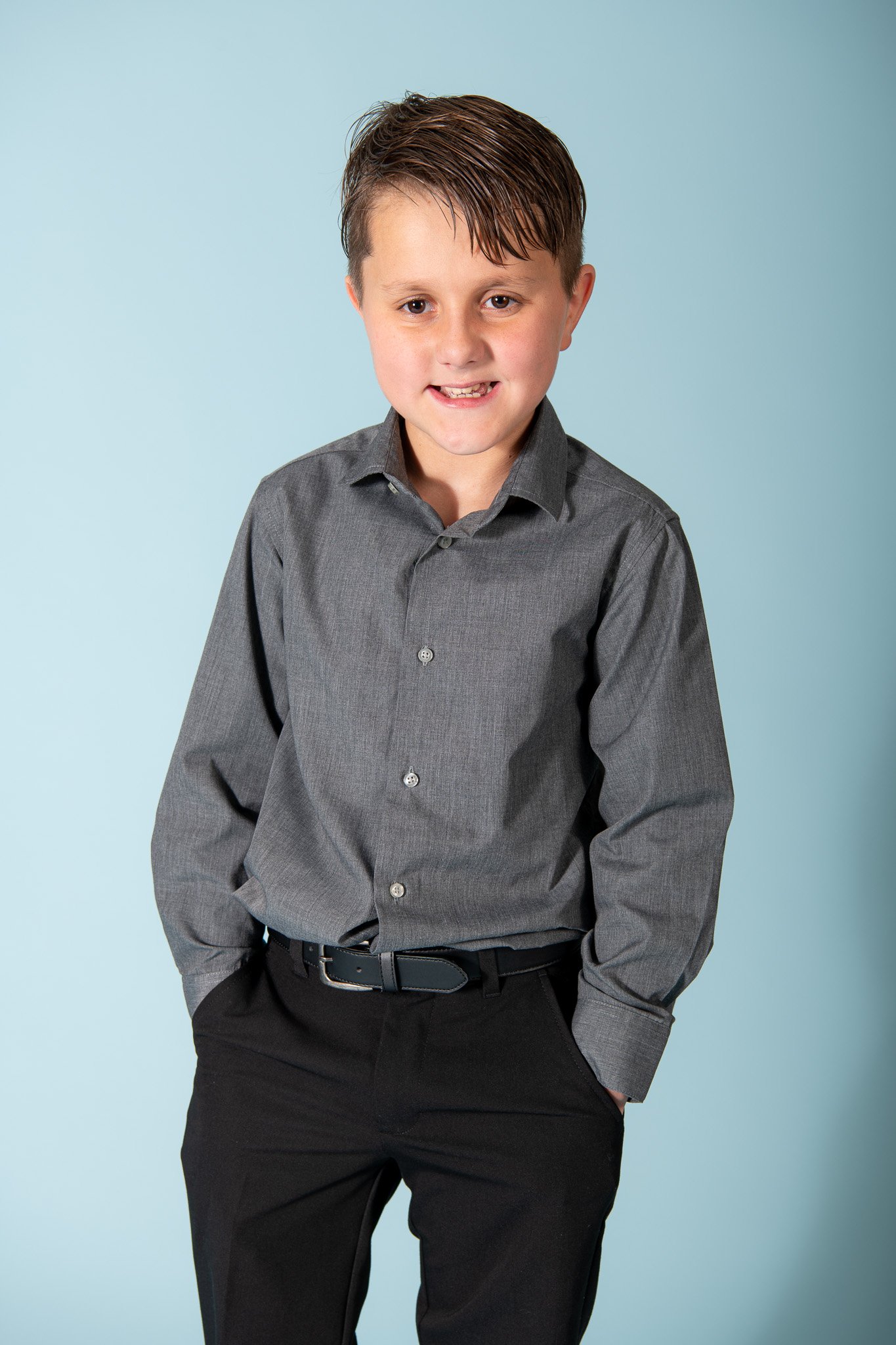 A young boy with brown hair, dressed in a gray shirt and black pants, standing against a light blue background.