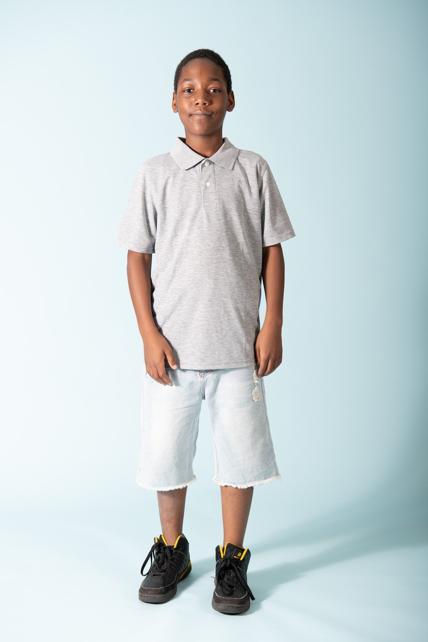 A boy standing against a light blue background, wearing a gray polo shirt, light denim shorts, and black sneakers with yellow accents.