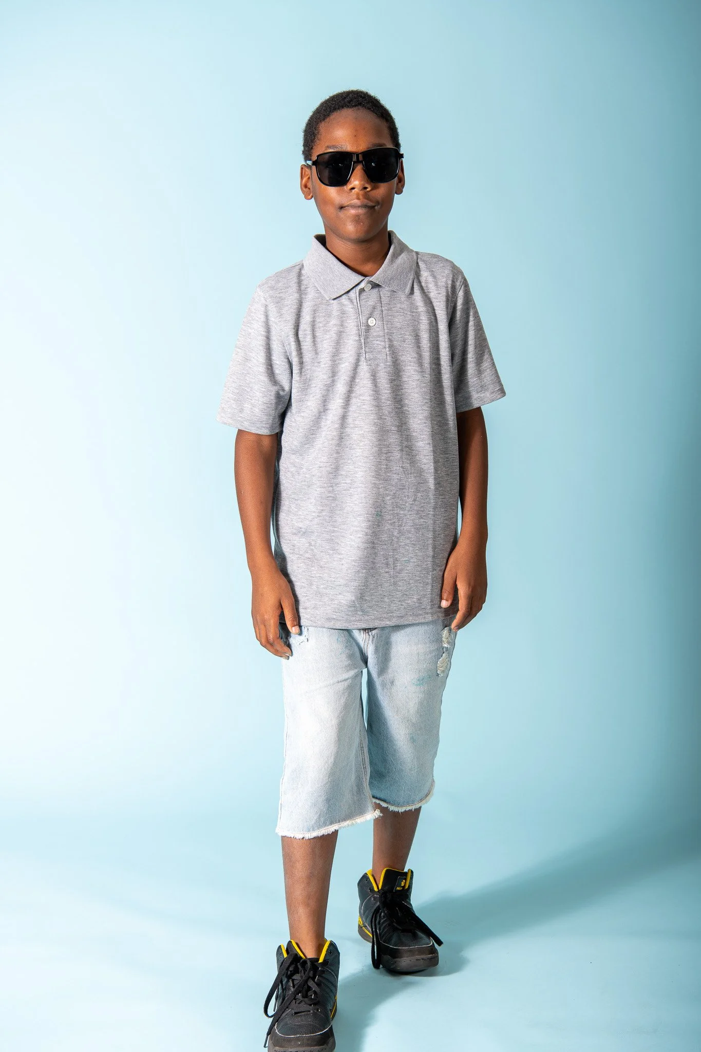 A boy wearing sunglasses, a gray polo shirt, ripped denim shorts, and black sneakers with yellow accents standing against a light blue background.