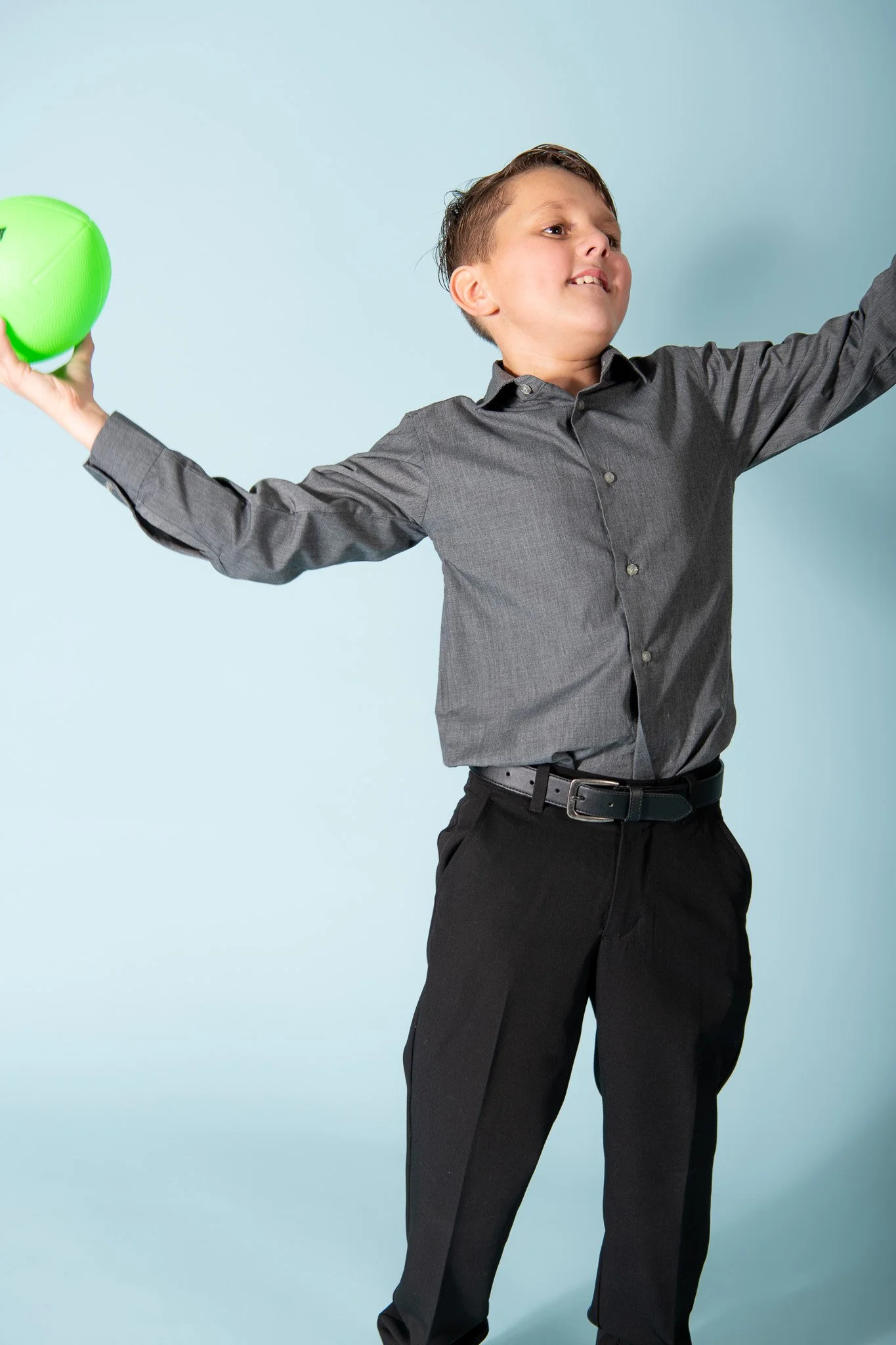 A young boy in a gray button-up shirt and black pants holding a bright green ball with arms outstretched.