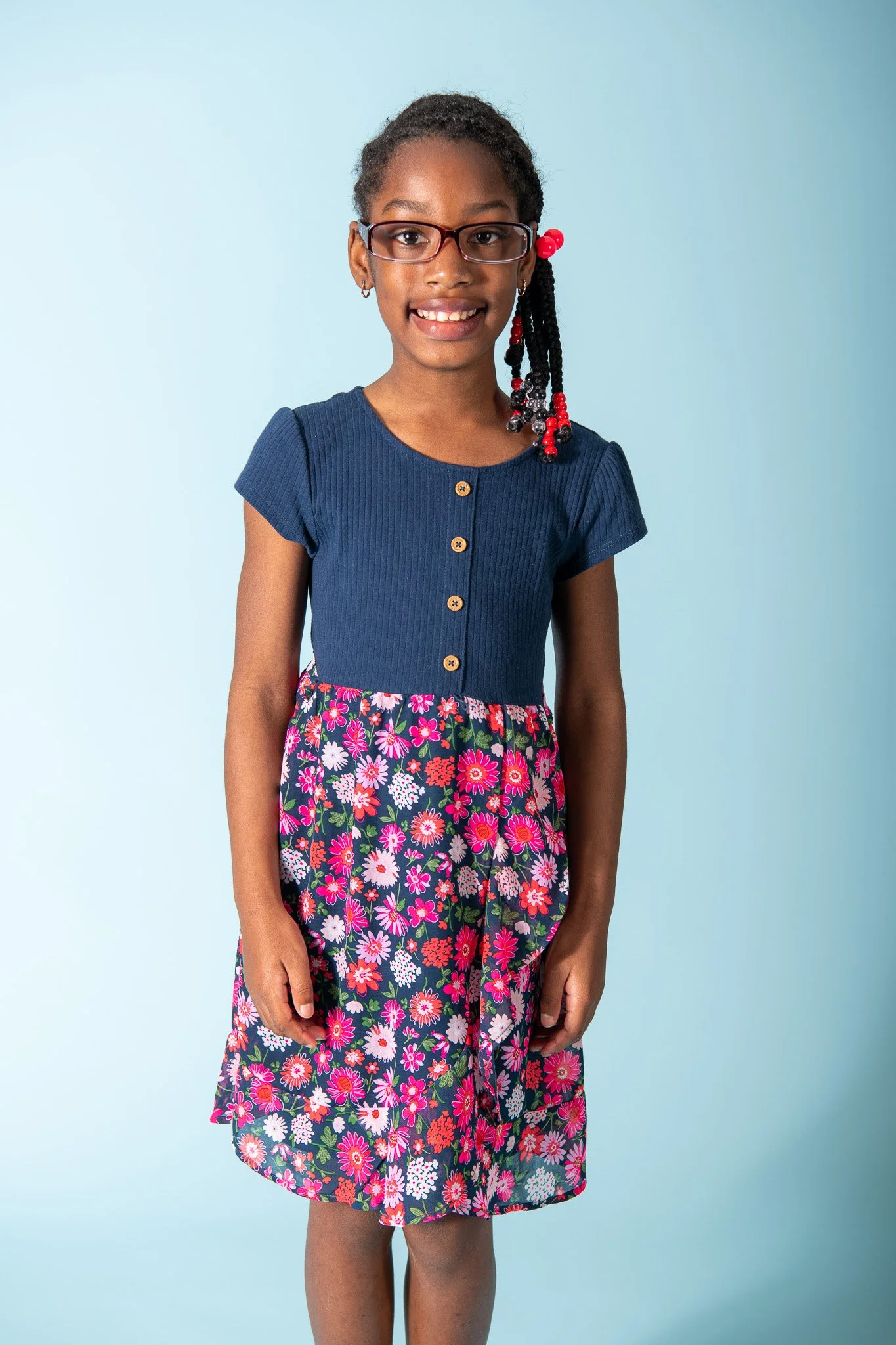 A young girl with glasses and braided hair decorated with red beads, wearing a blue top with buttons and a floral skirt, standing against a light blue background.