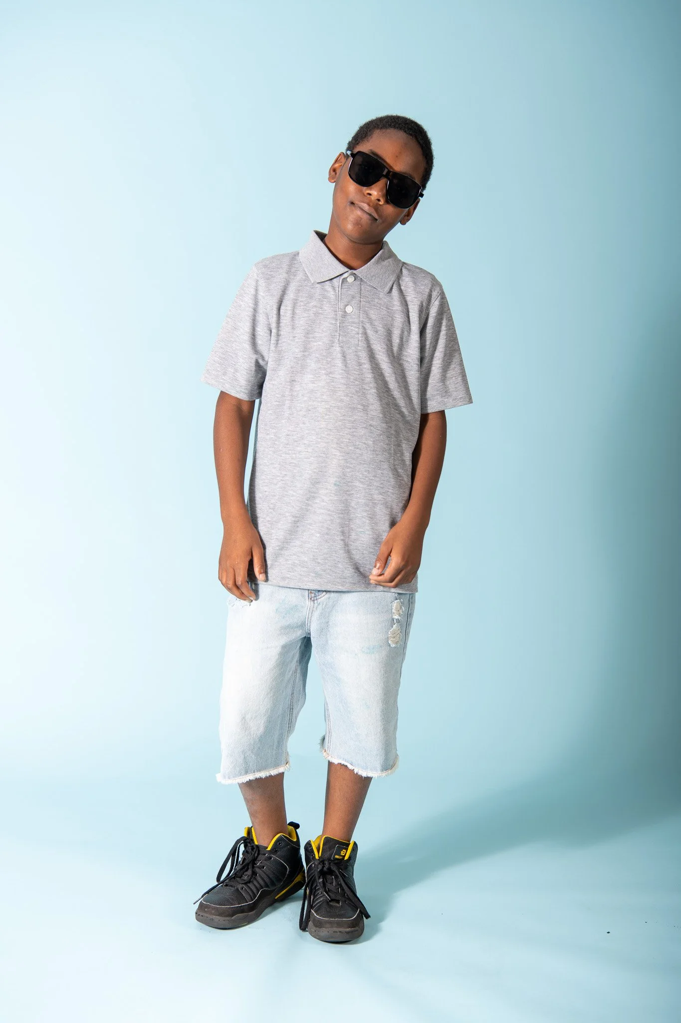 A young boy wearing a gray polo shirt, distressed denim shorts, black sneakers with yellow accents, and black sunglasses, standing against a light blue background.