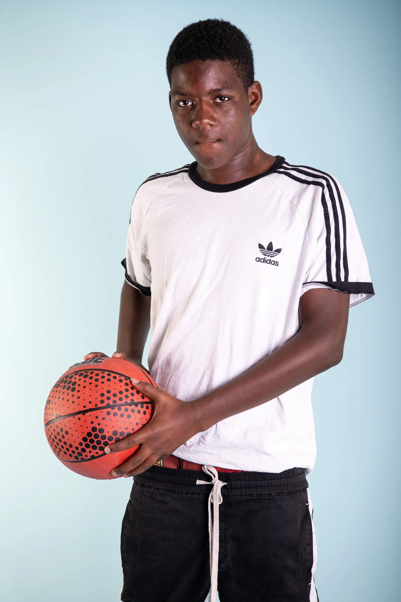 A young man holding a basketball, wearing a white Adidas t-shirt and black shorts, against a light blue background.