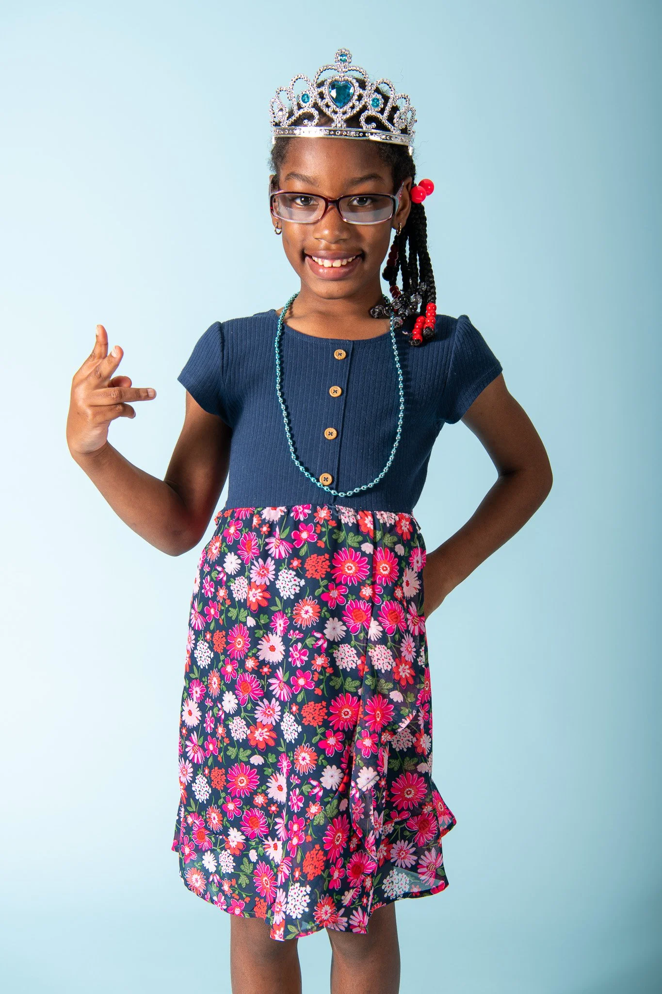 Young girl wearing a crown, glasses, a navy blue top, and a floral skirt, striking a confident pose with smile and peace sign.
