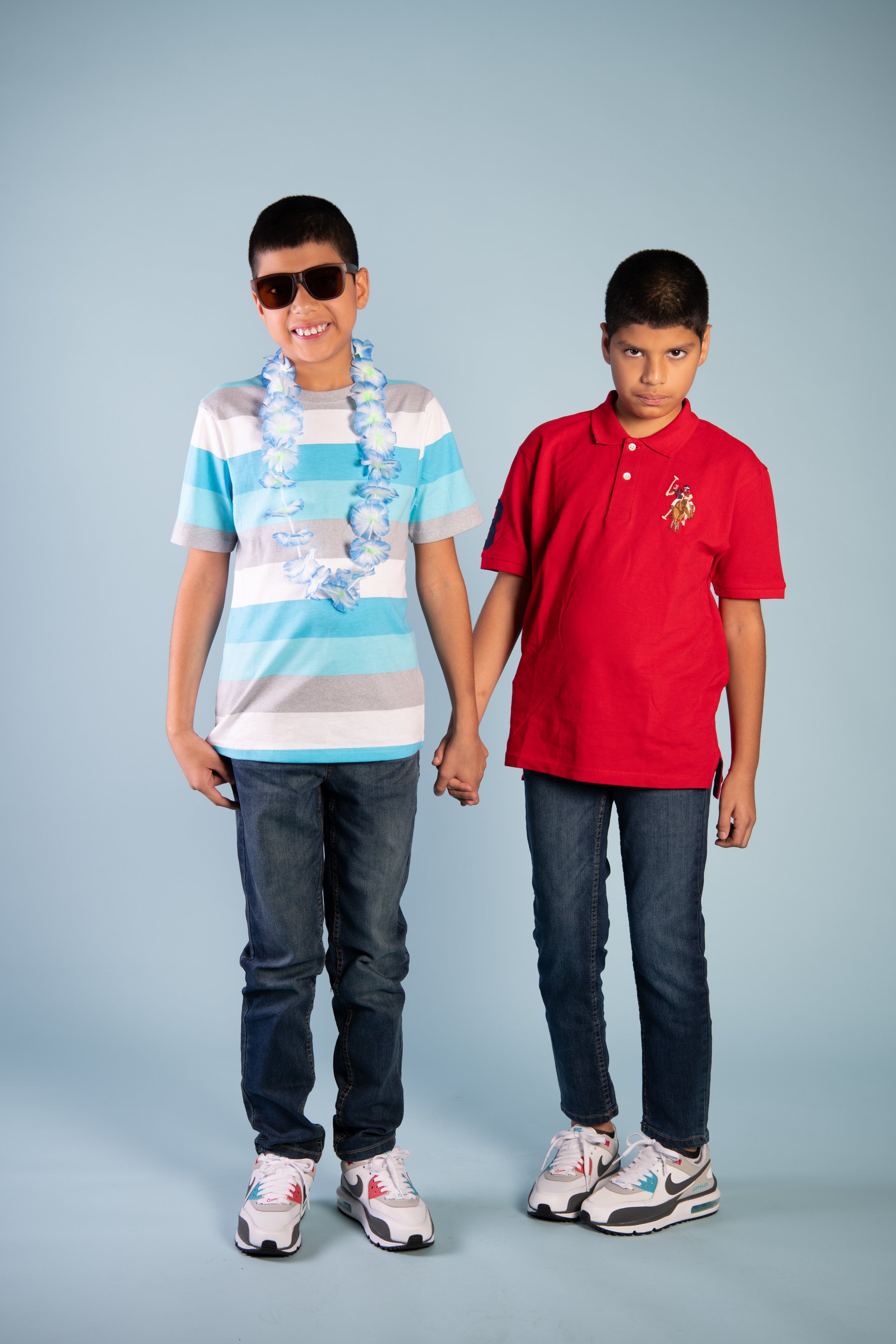 Two young boys holding hands, one with sunglasses and a flower lei, dressed casually in jeans and sneakers, standing against a plain background.
